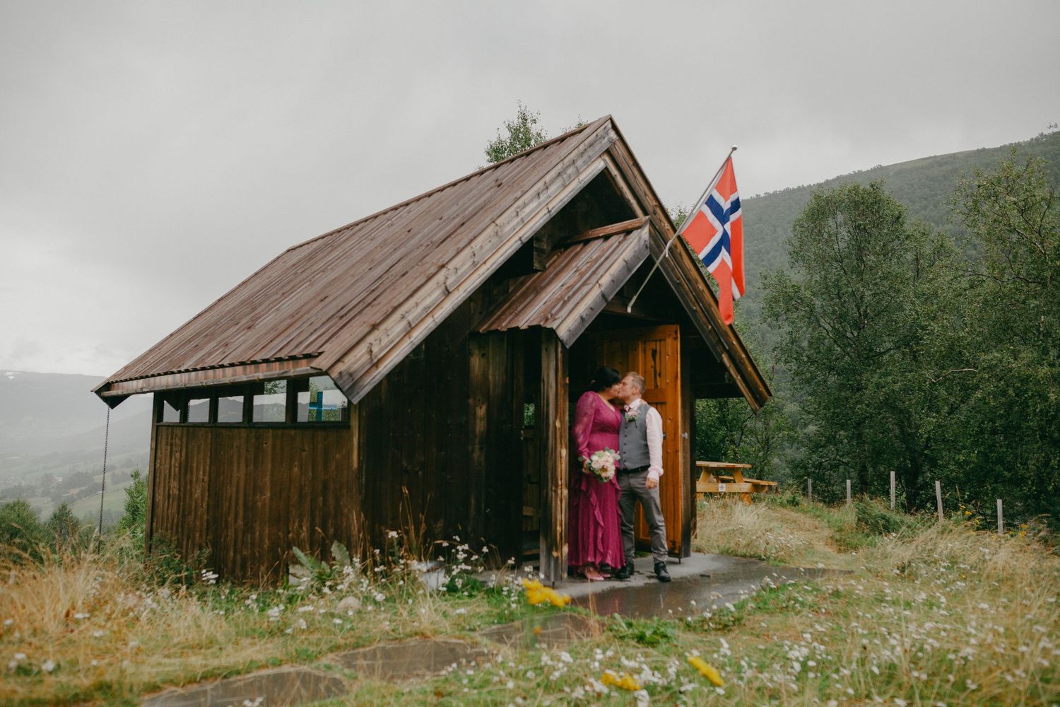 Couple embracing at a wooden cabin with Norwegian flag in a lush green mountain landscape on a cloudy day.