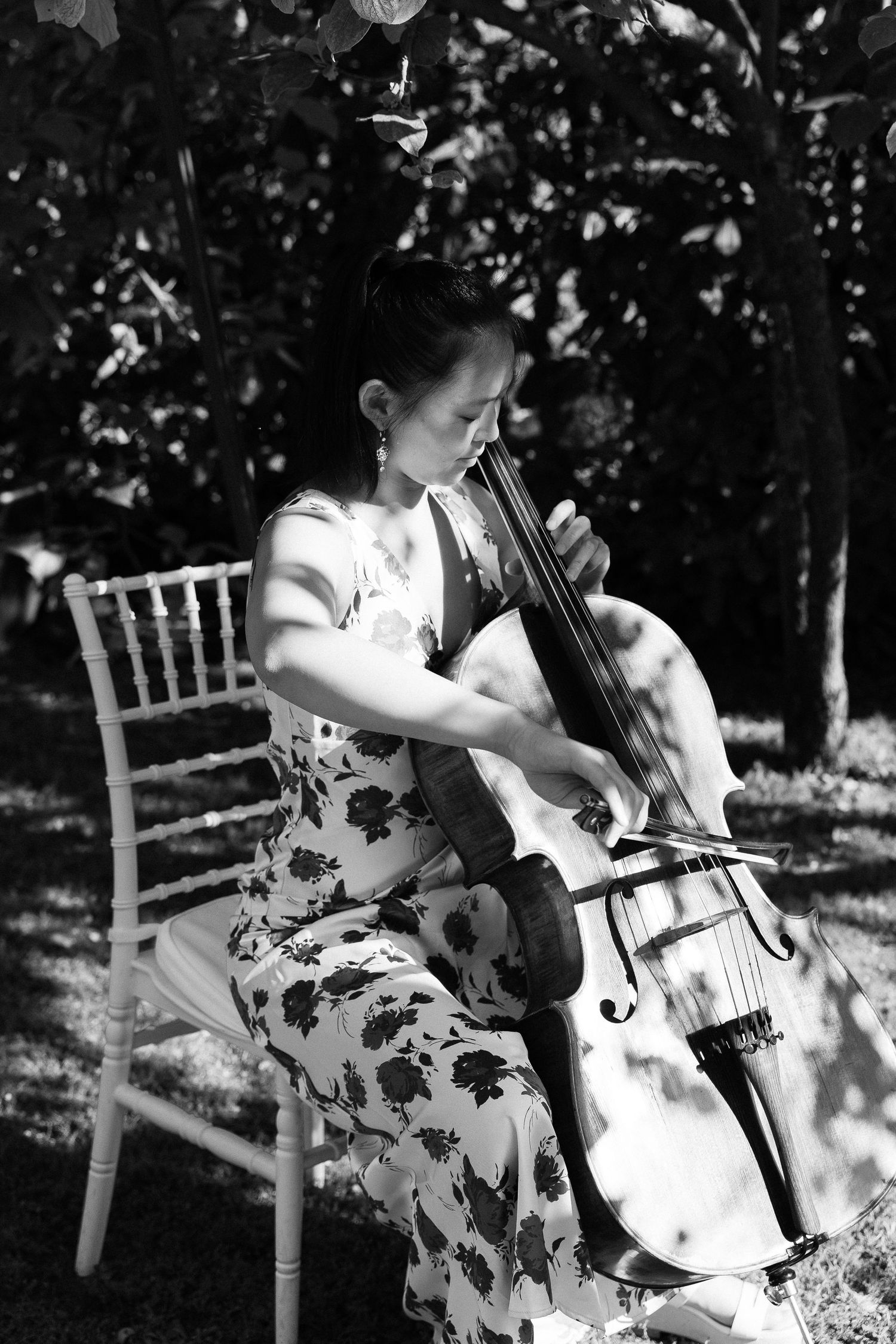 Black and white photo of woman playing cello outdoors in floral dress at wedding ceremony.