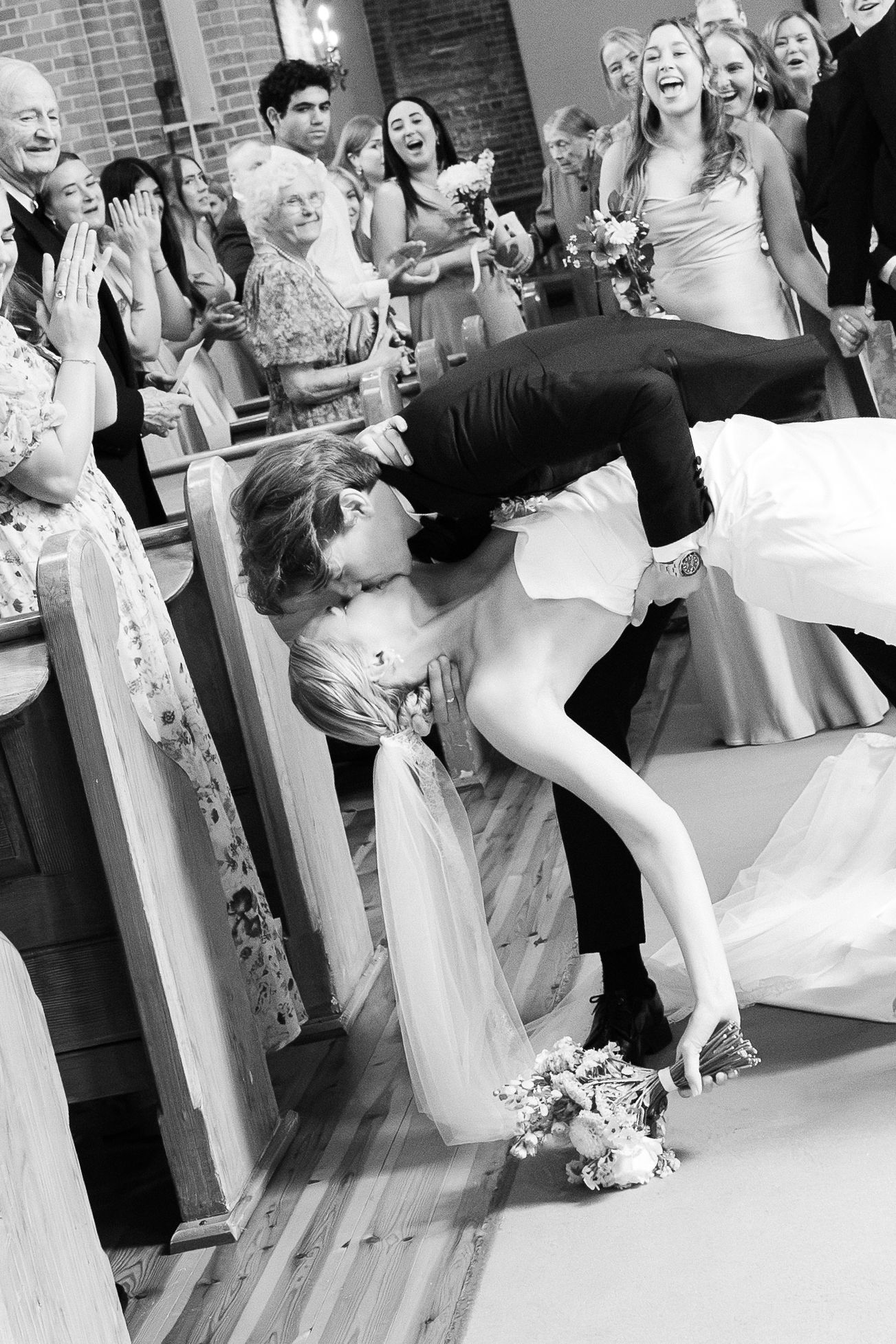Black and white photo of a groom dipping his bride for a kiss at their wedding ceremony while guests cheer.