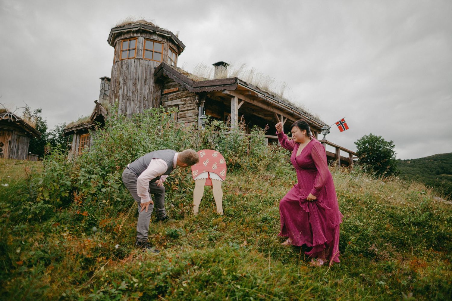 Family playing outdoors near a rustic wooden building with grass roof on a cloudy day in Norway.