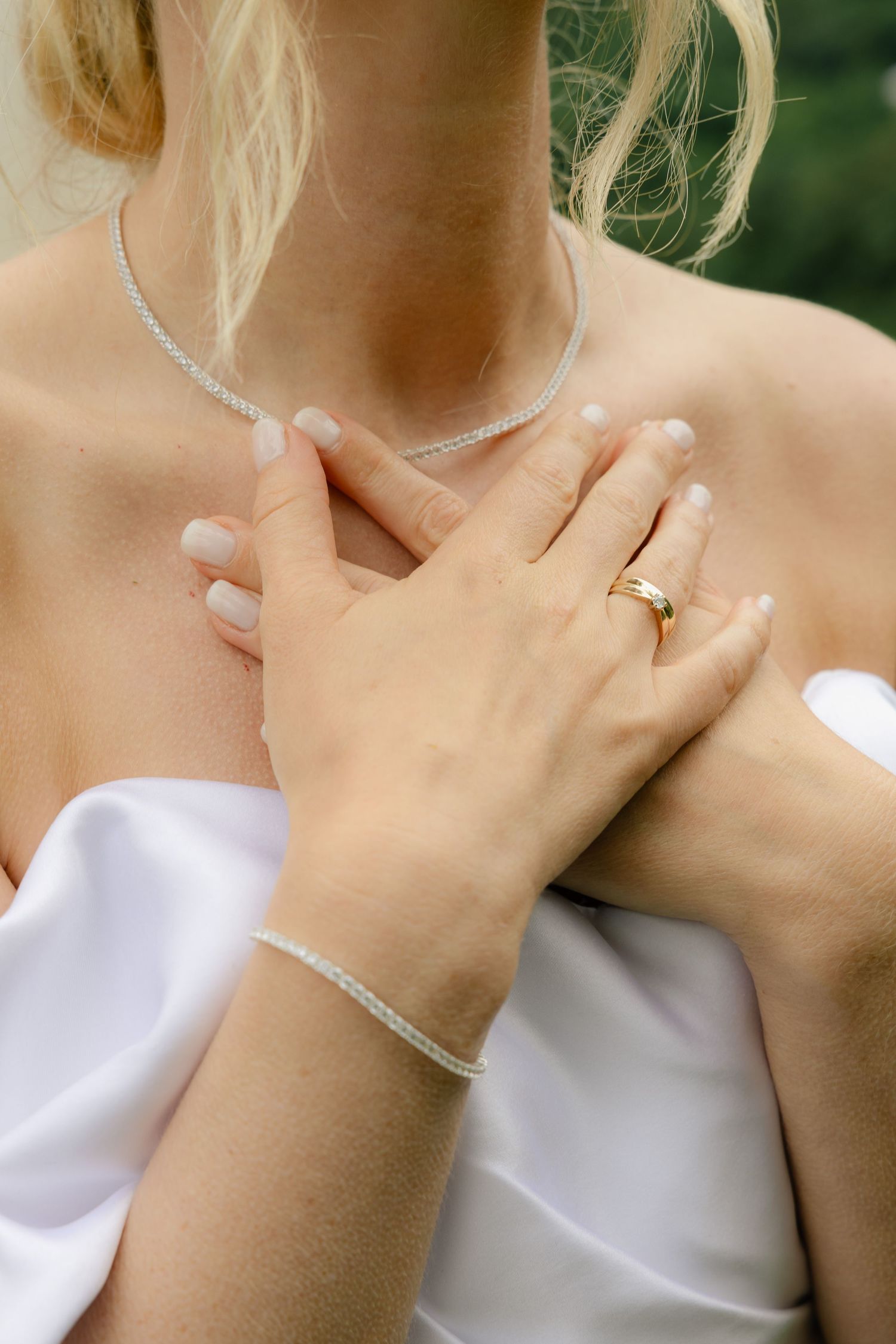 A bride in a white off-shoulder gown displays her gold wedding ring, bracelet, and diamond necklace.