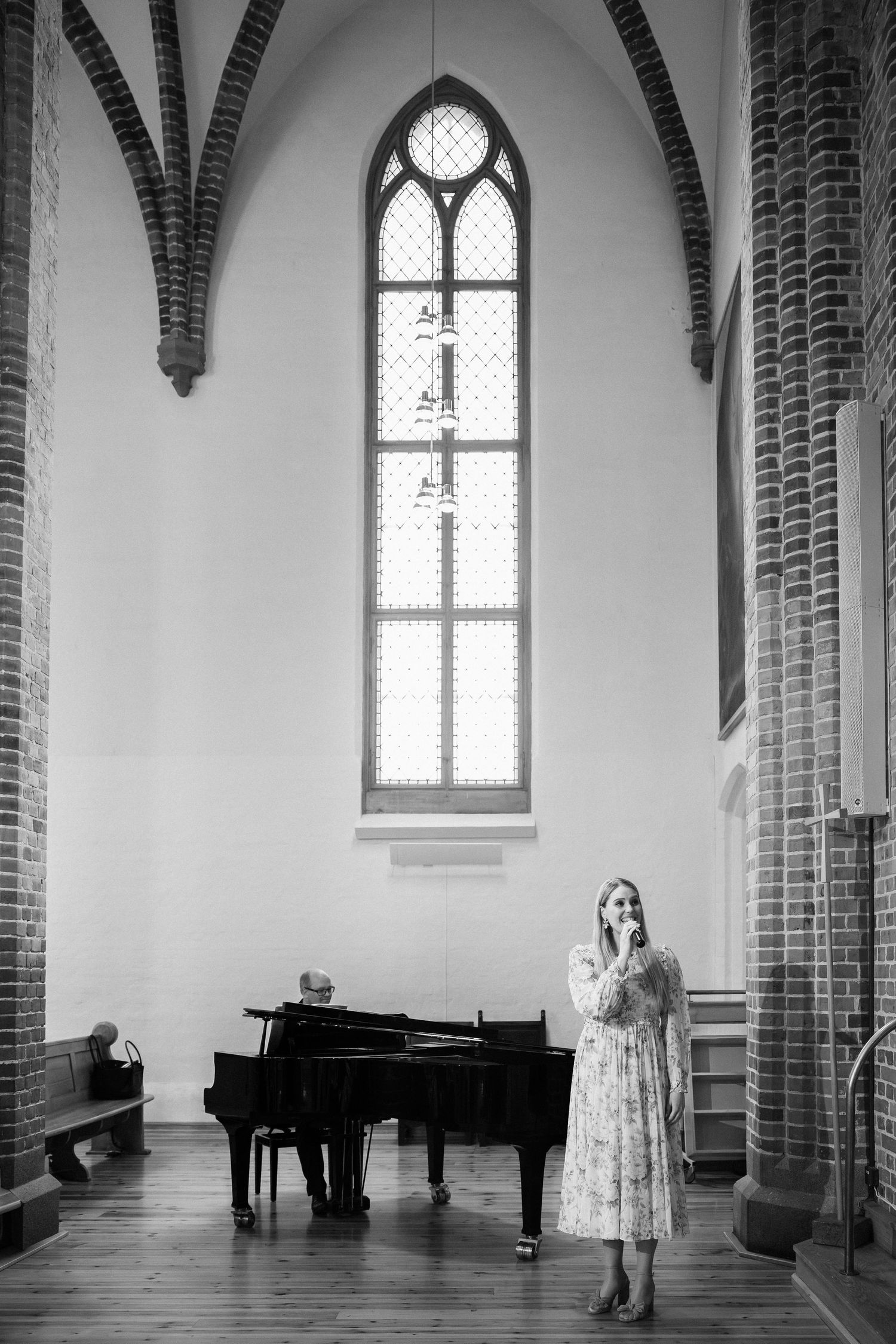 Black and white photo of woman in floral dress standing in church with grand piano and arched window.