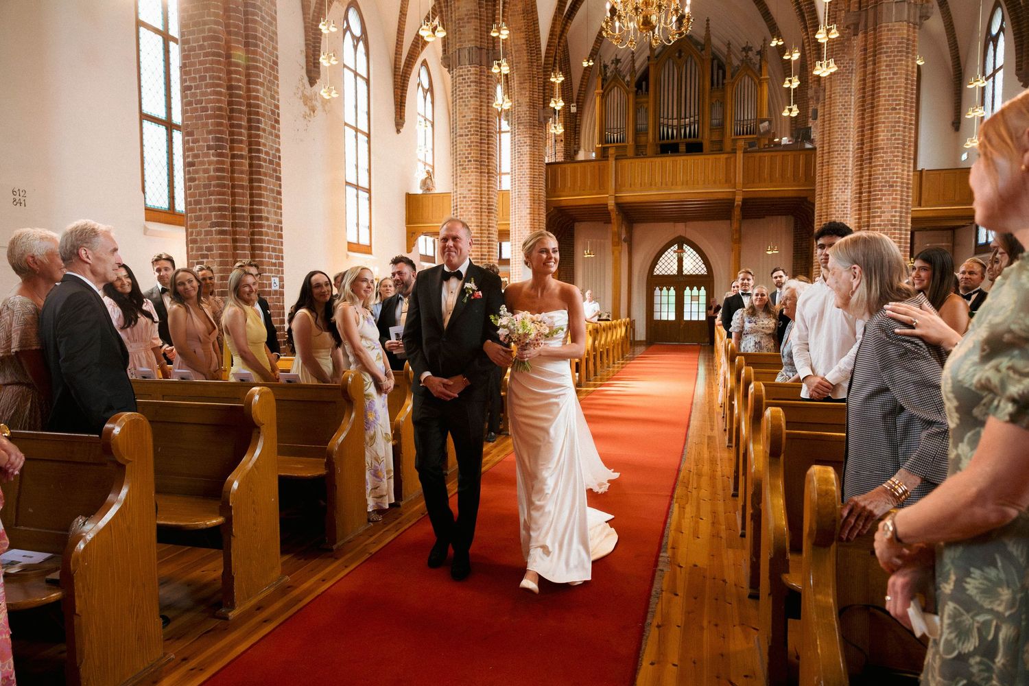 Bride and groom walk down red carpet aisle in elegant brick church surrounded by smiling wedding guests.