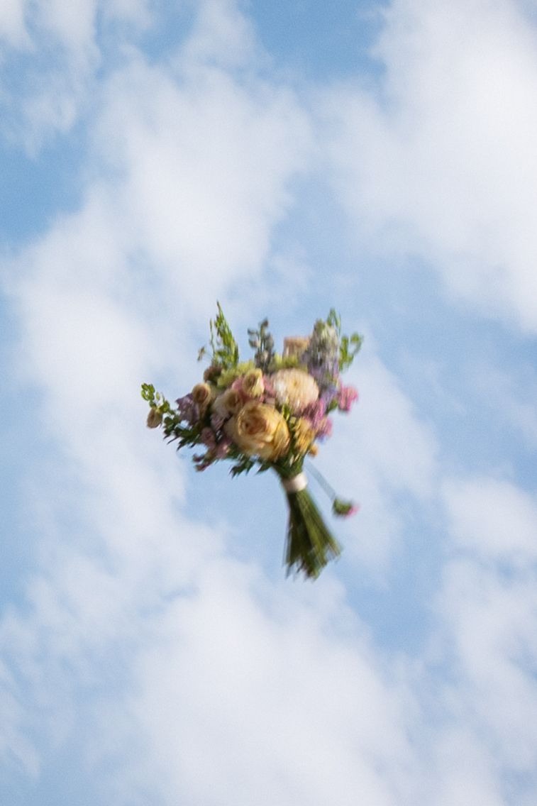 A colorful wedding bouquet tossed in the air against a blue sky with white clouds.