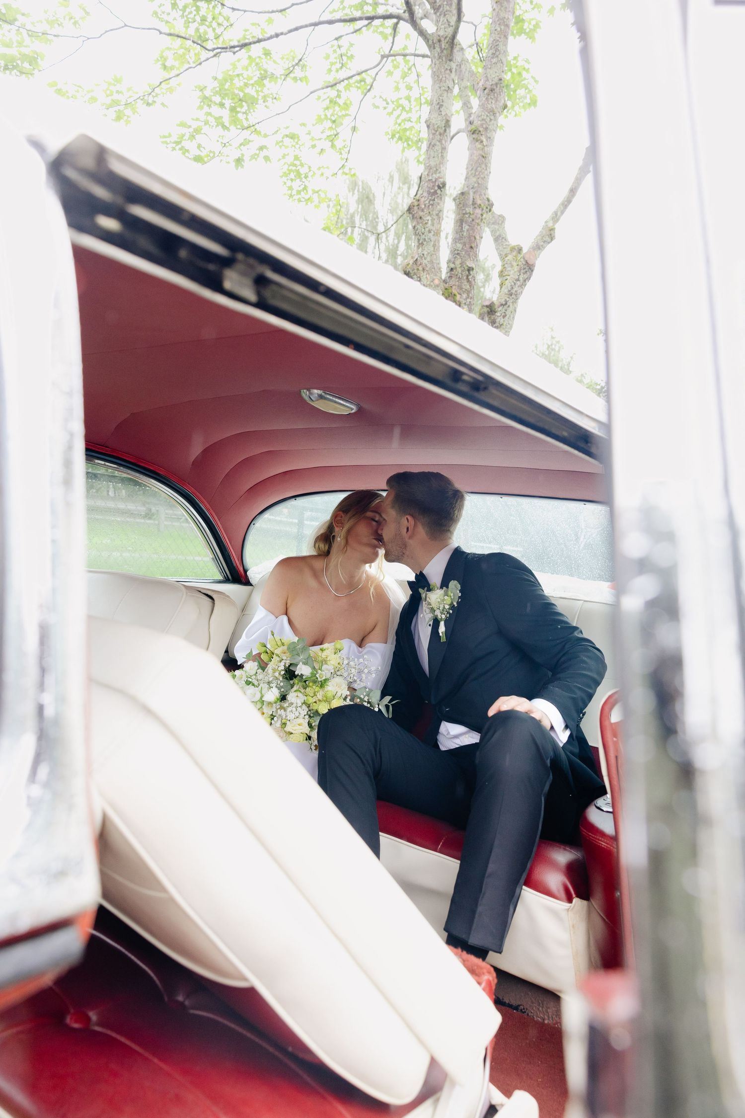 Newlyweds sharing a romantic kiss in the backseat of a classic vintage red car.