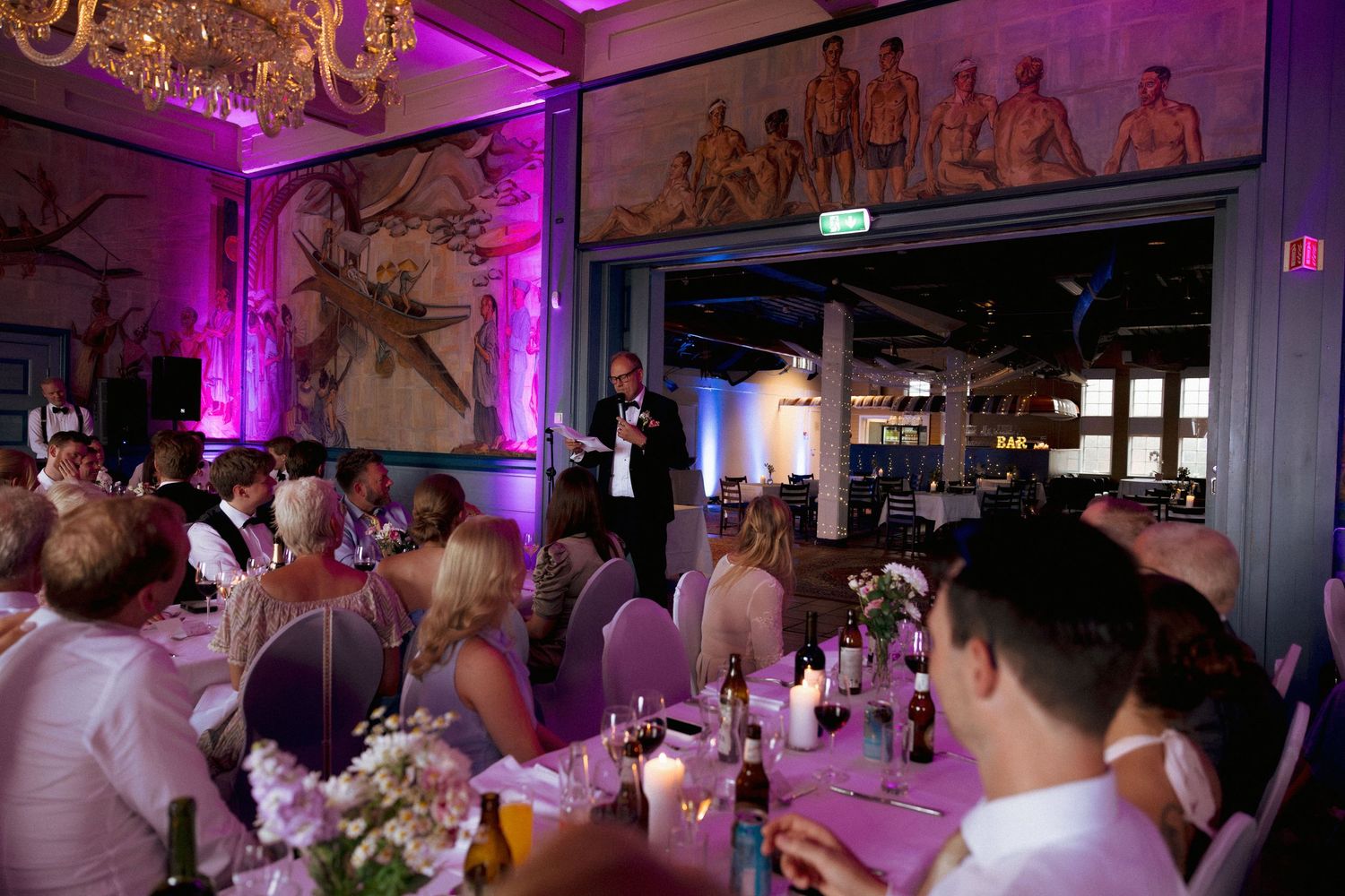 Wedding reception dinner with guests seated at decorated tables while man gives speech in purple-lit venue.