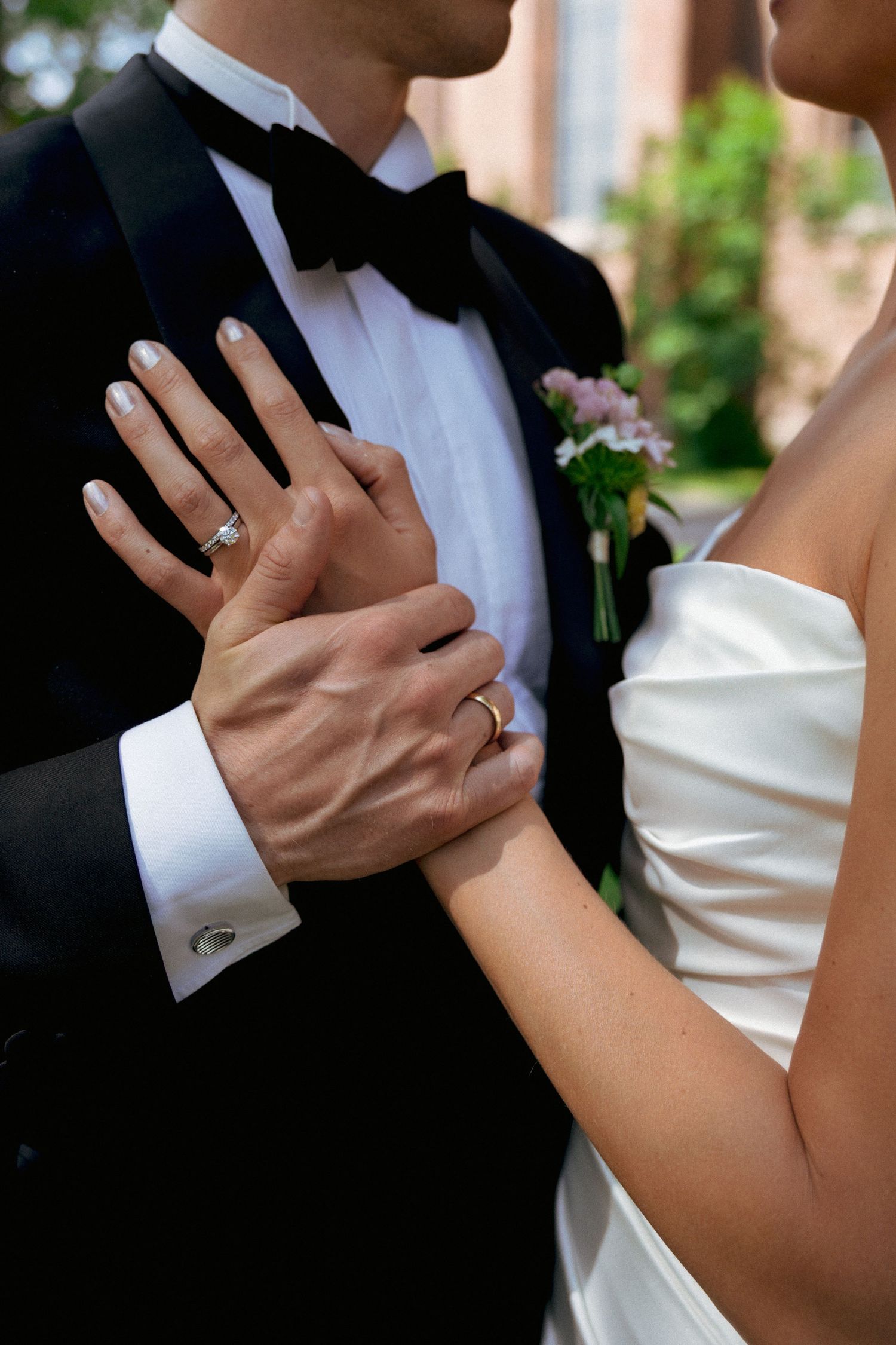 Close-up of bride and groom holding hands showing wedding rings, groom in tuxedo with boutonniere.