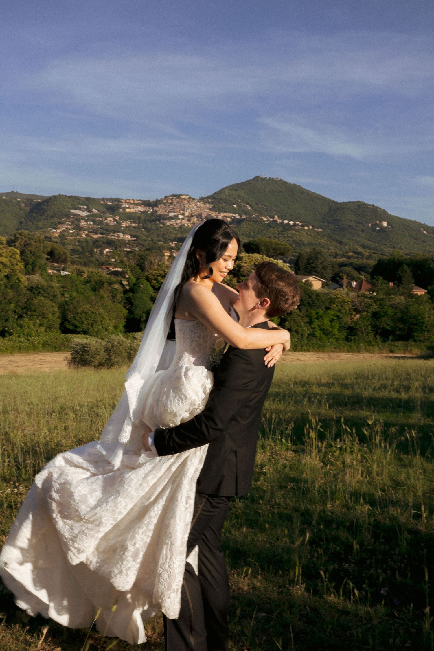 Groom lifts bride in white lace gown in sunlit field with mountain landscape backdrop.