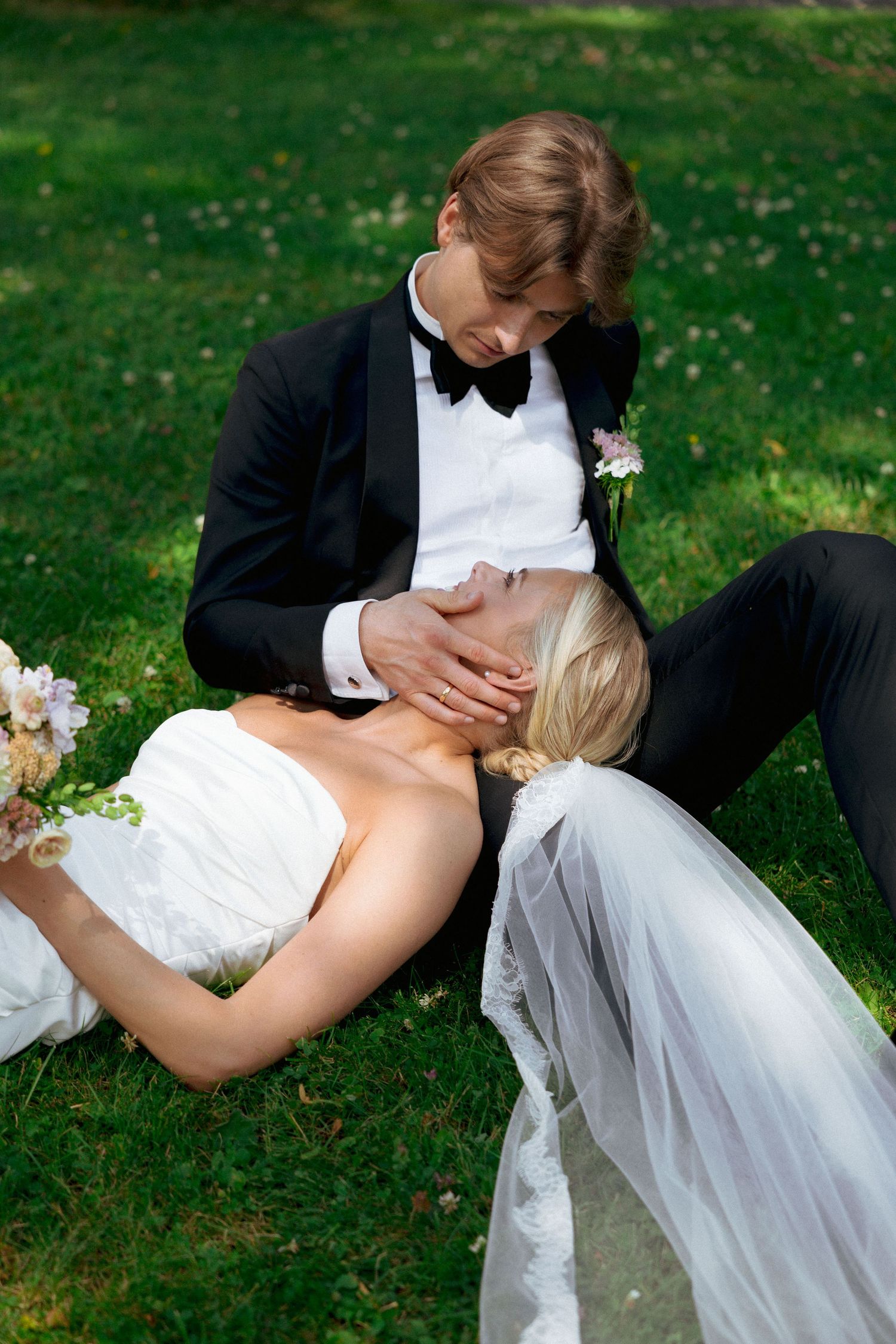 Bride in white gown rests her head on groom's lap while sitting on green grass during outdoor wedding photo session.