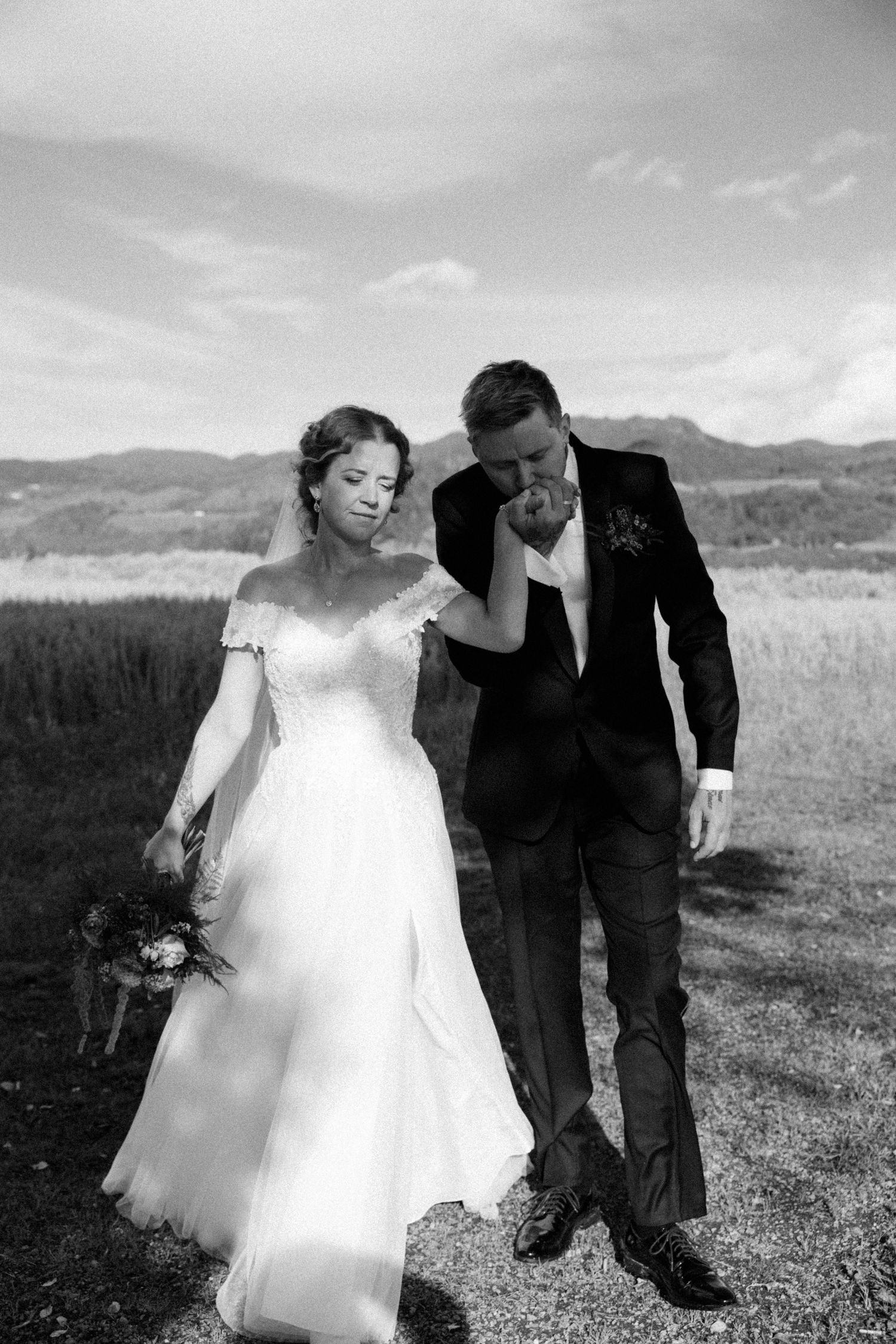 Black and white photo of a groom kissing his bride's hand outdoors with mountains in the background.