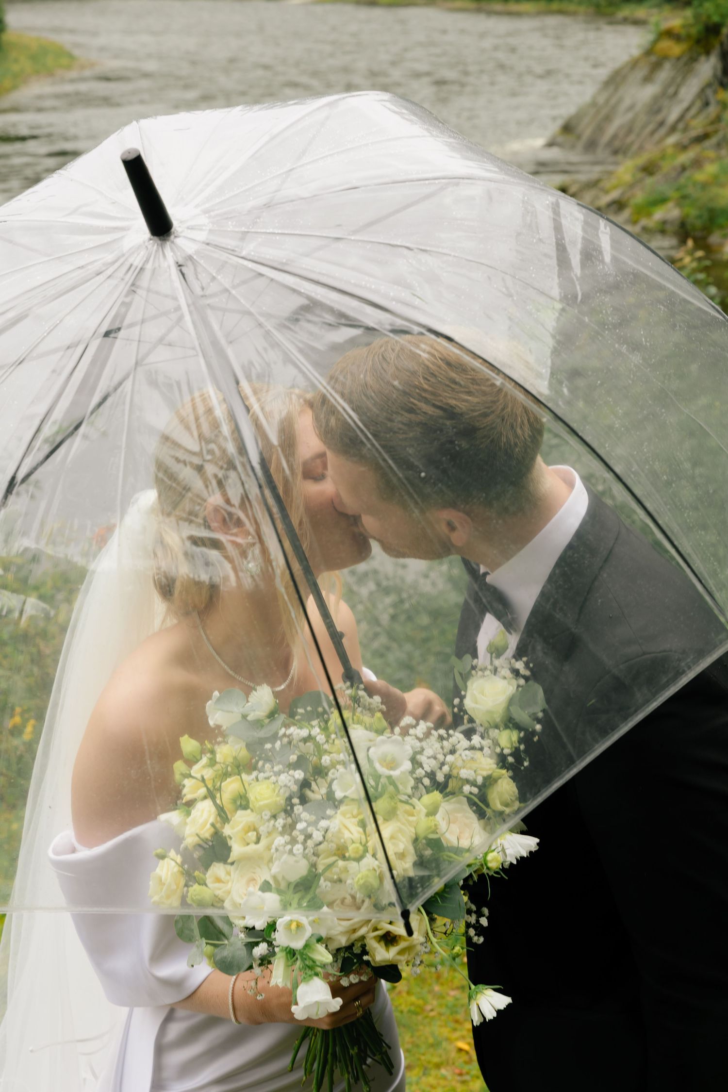 Bride and groom share a romantic kiss under a clear umbrella while holding a white floral bouquet in the rain.