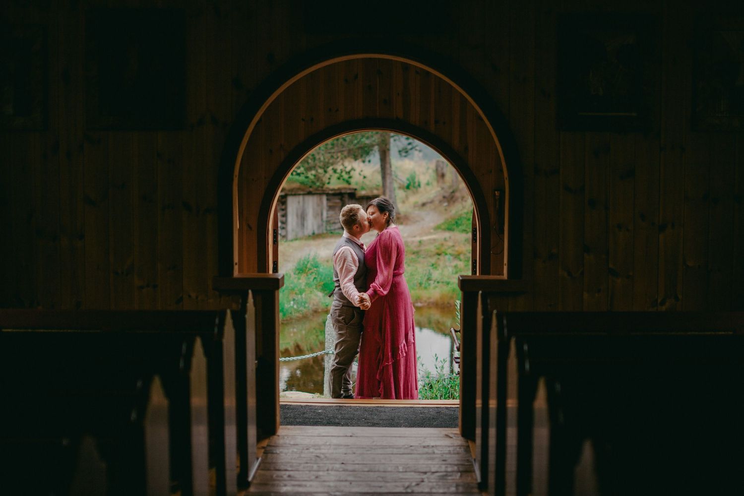 Couple embracing in a rustic arched doorway with lush greenery and water visible in the background.