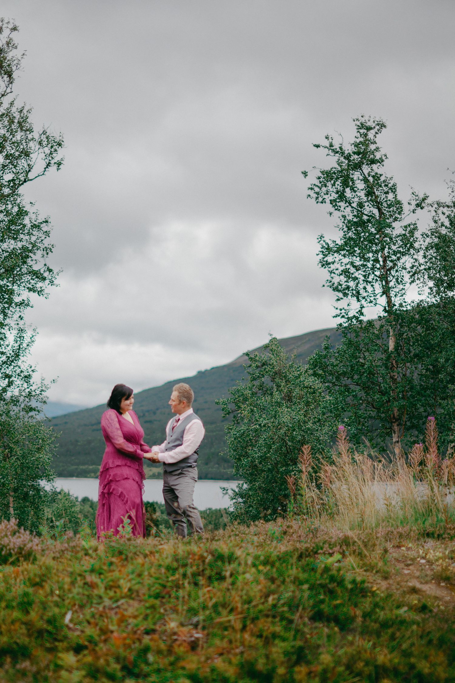 Couple holding hands in a lush green landscape with a lake and mountains under cloudy skies.
