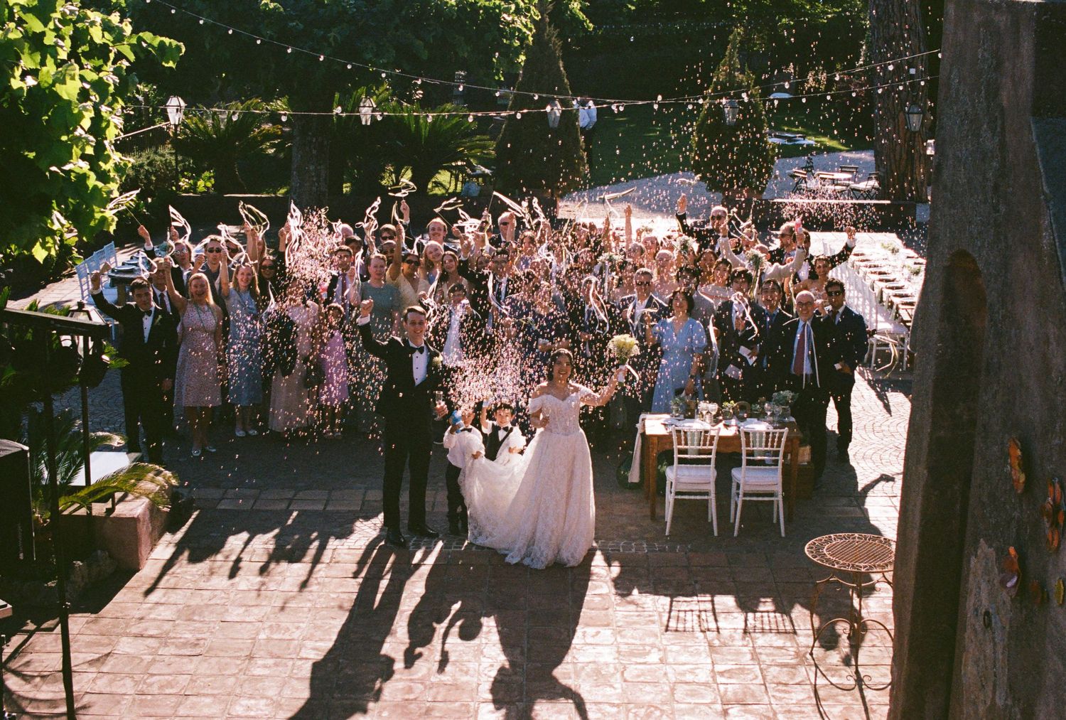 Bride and groom celebrate with cheering wedding guests outdoors at a sunlit garden reception venue.