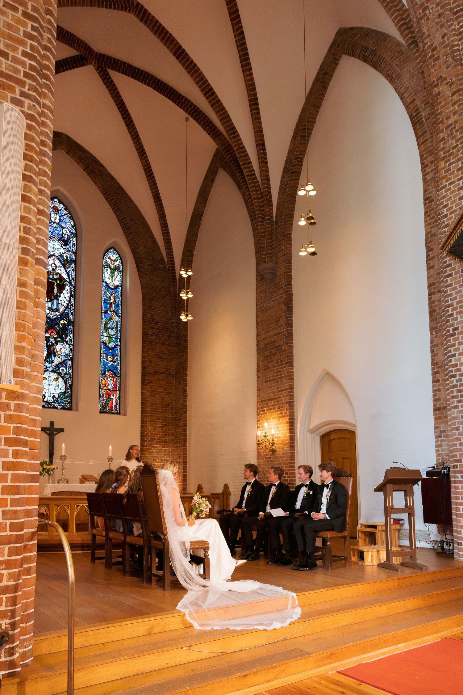 Wedding ceremony inside a brick Gothic church with stained glass windows, groomsmen seated in pews, and bride's dress draped over a chair.
