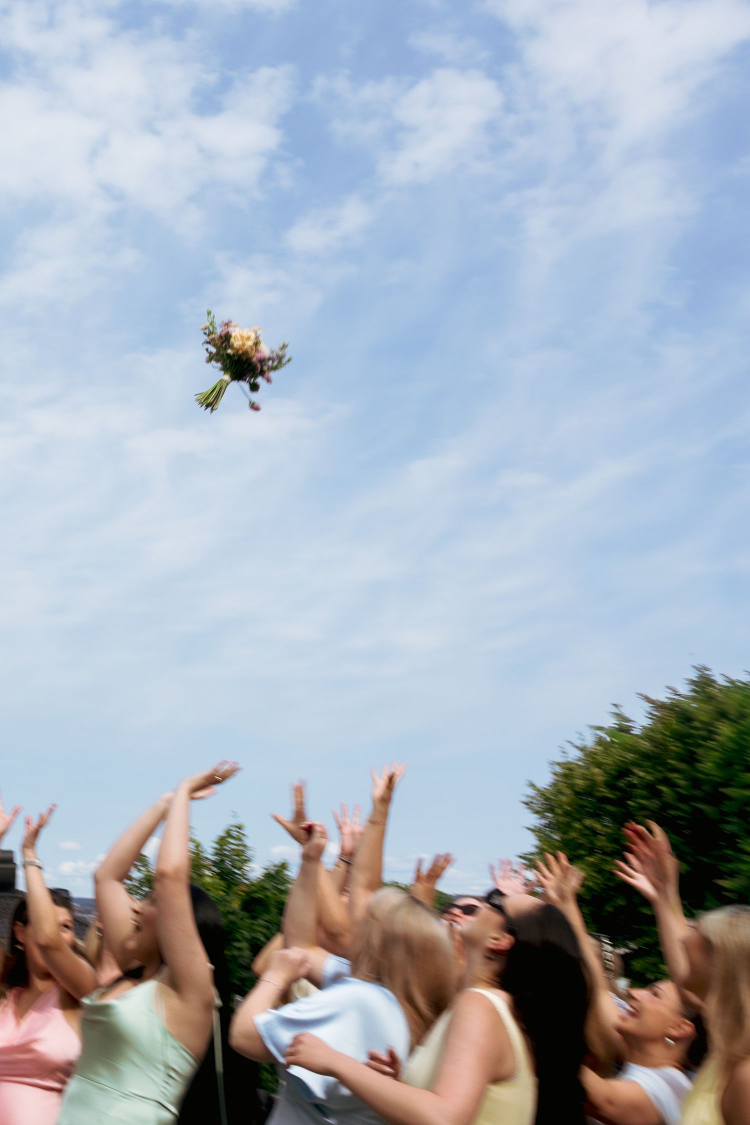 Excited wedding guests reaching up to catch the brides bouquet tossed into a blue sky.