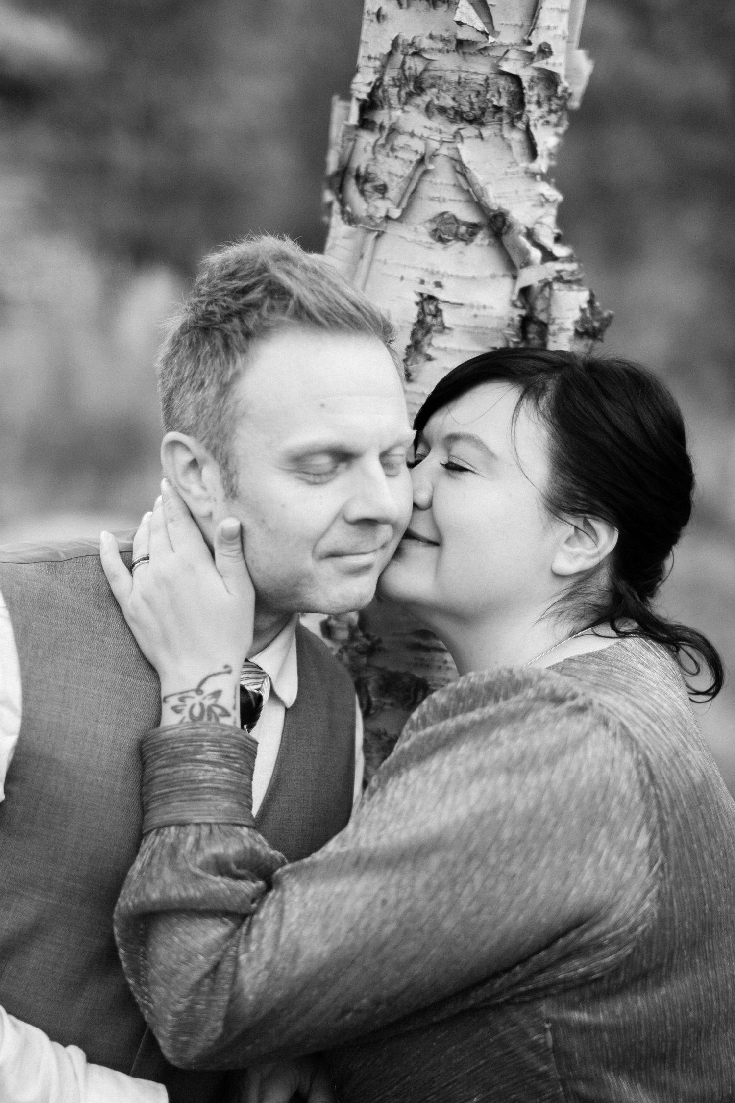 Black and white photo of a woman kissing a mans cheek tenderly near a birch tree outdoors.