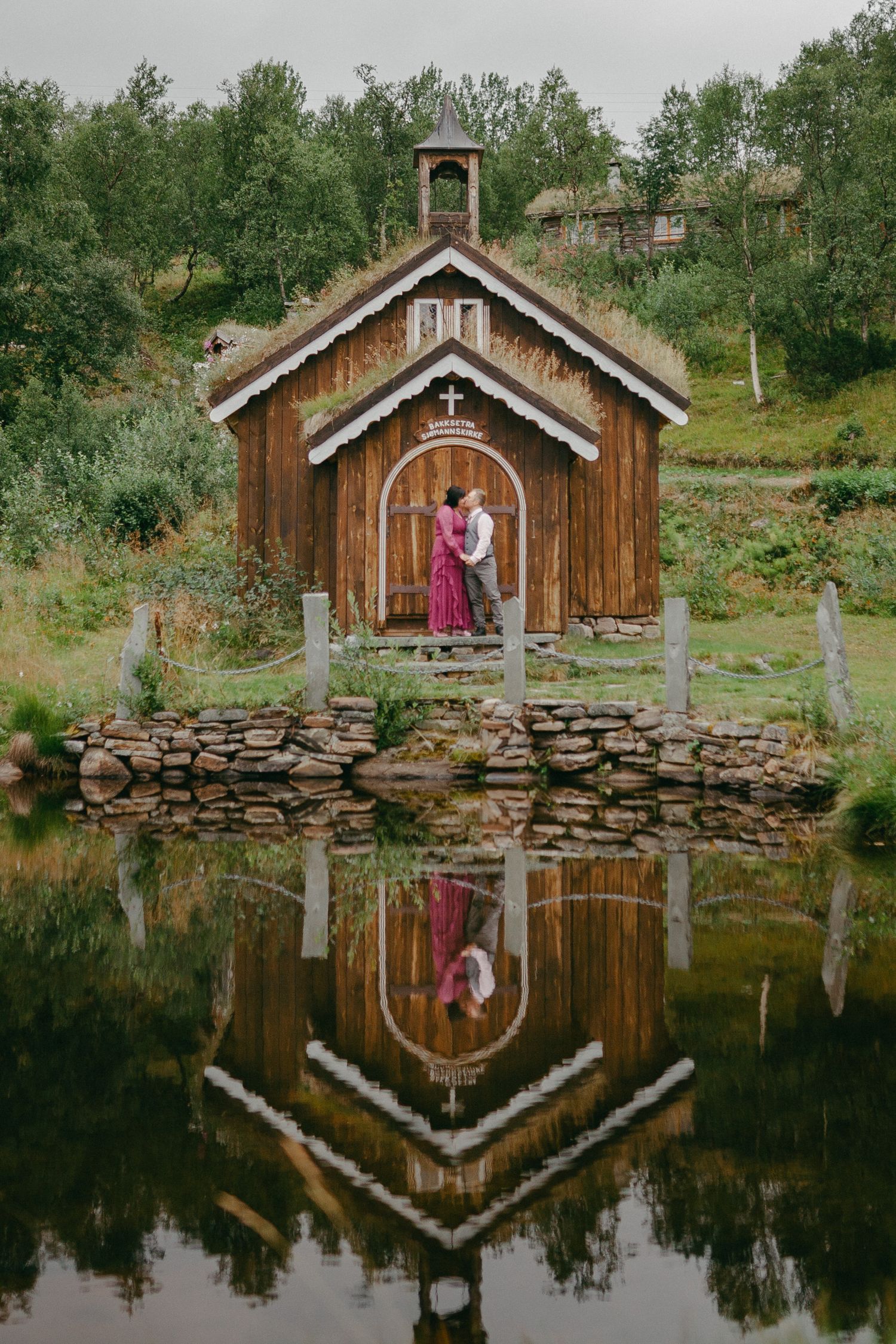 Couple embracing before a rustic wooden chapel with a cross, reflected in a still pond, surrounded by lush greenery.