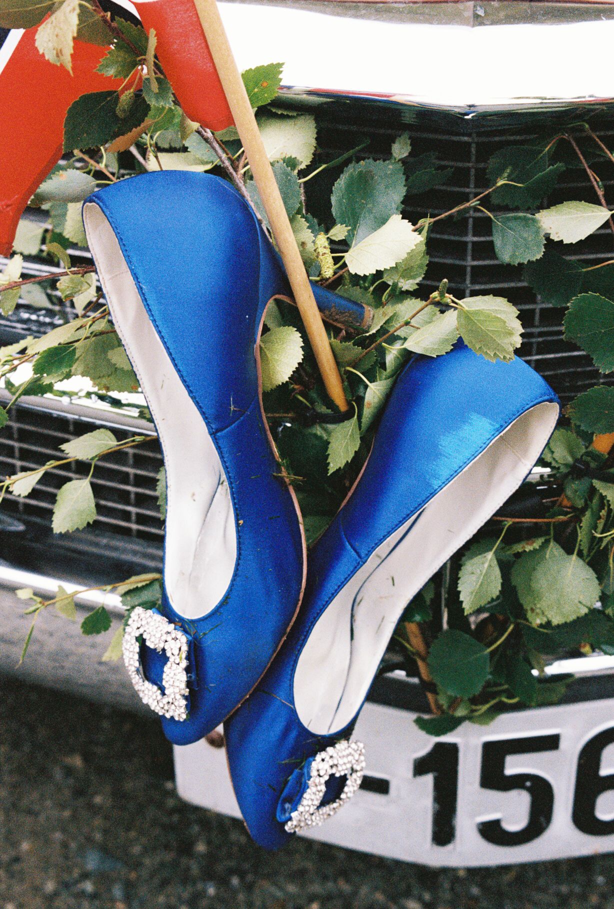 Elegant blue satin heels with crystal buckles displayed on a vintage car grille with green leaves.
