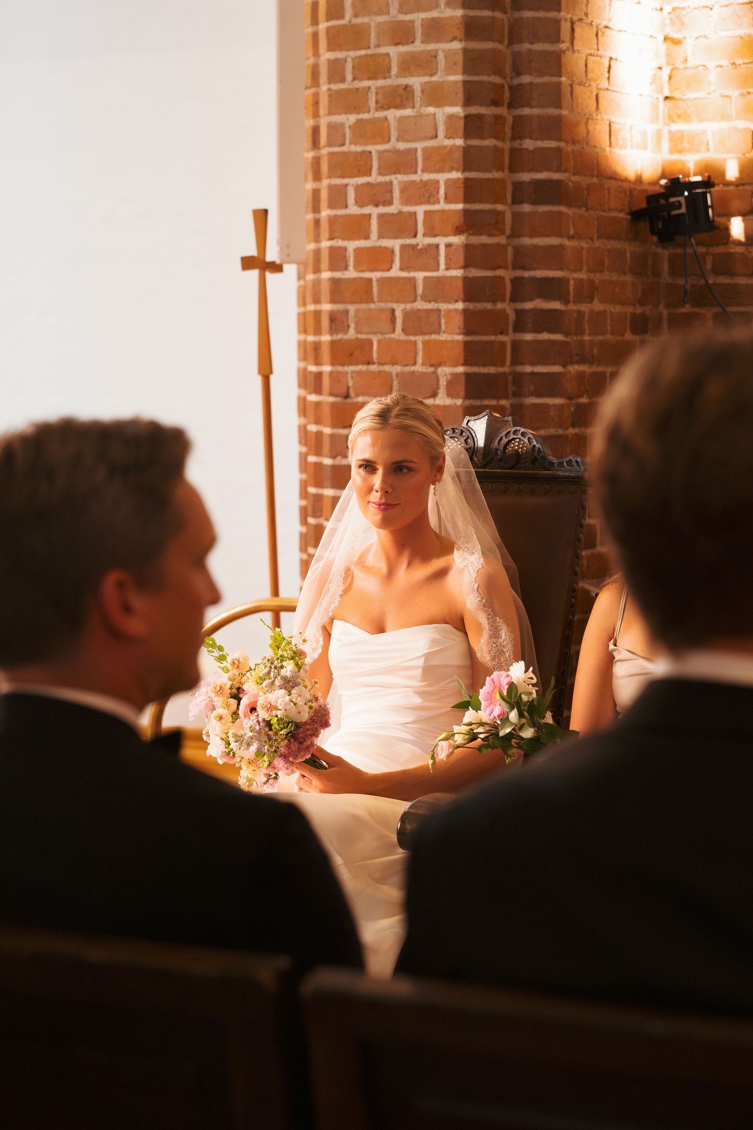 Bride in white strapless gown and veil holds pink bouquet while seated at wedding ceremony in brick church.