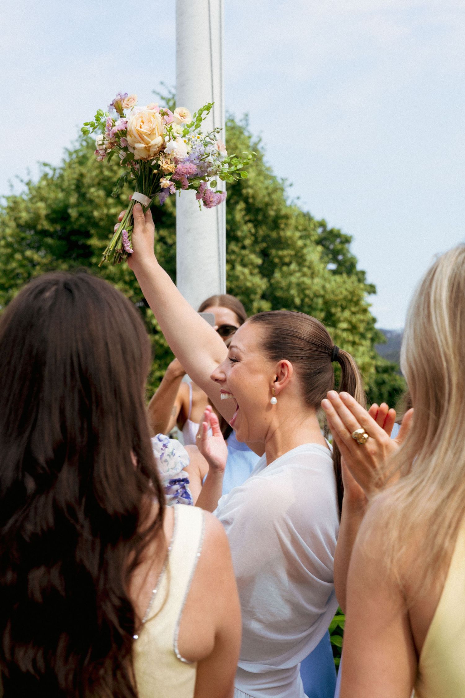 Excited woman catches bridal bouquet toss surrounded by cheering guests at outdoor wedding reception.