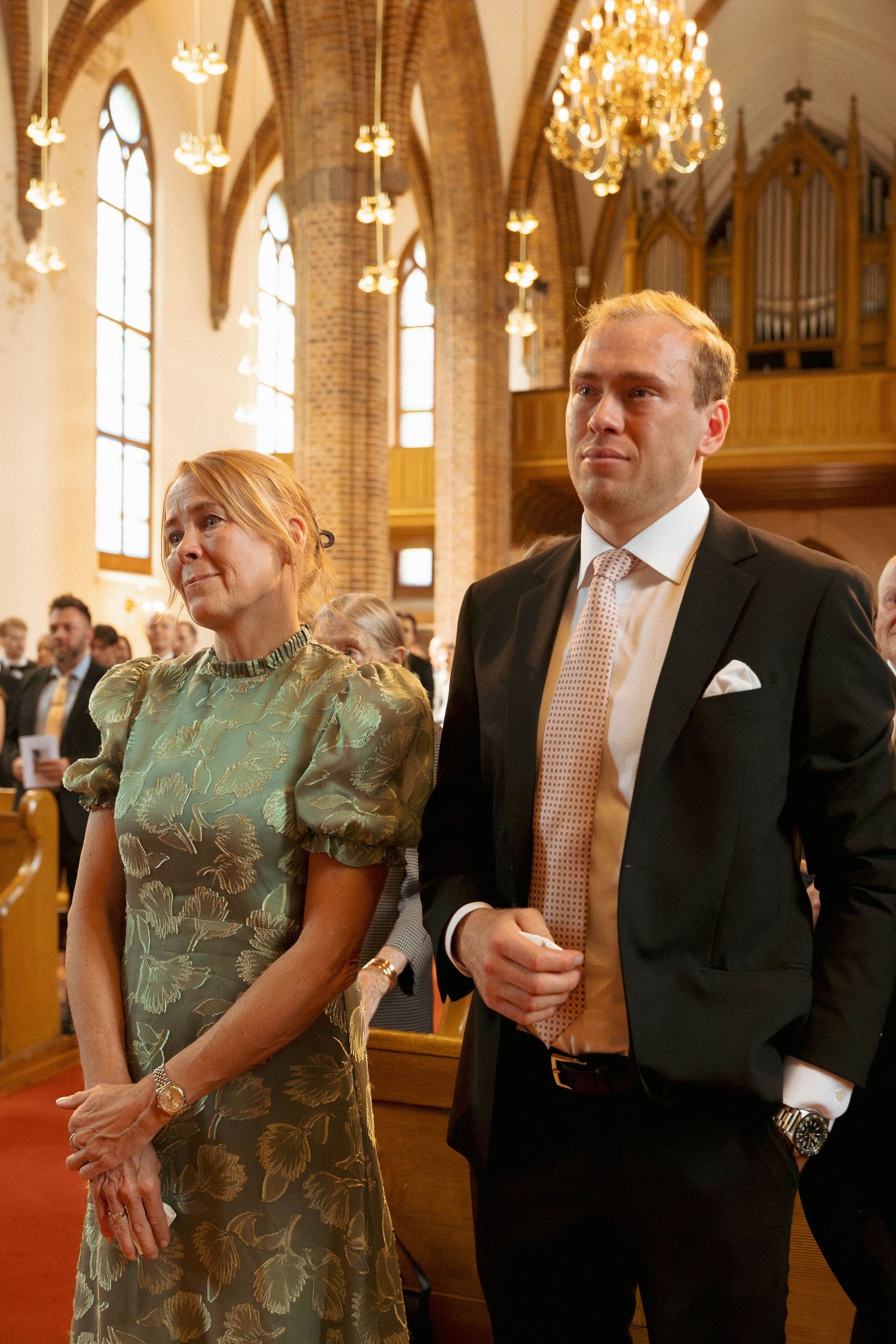 Elegant couple in formal attire standing inside a church with chandeliers, woman in green floral dress, man in dark suit.