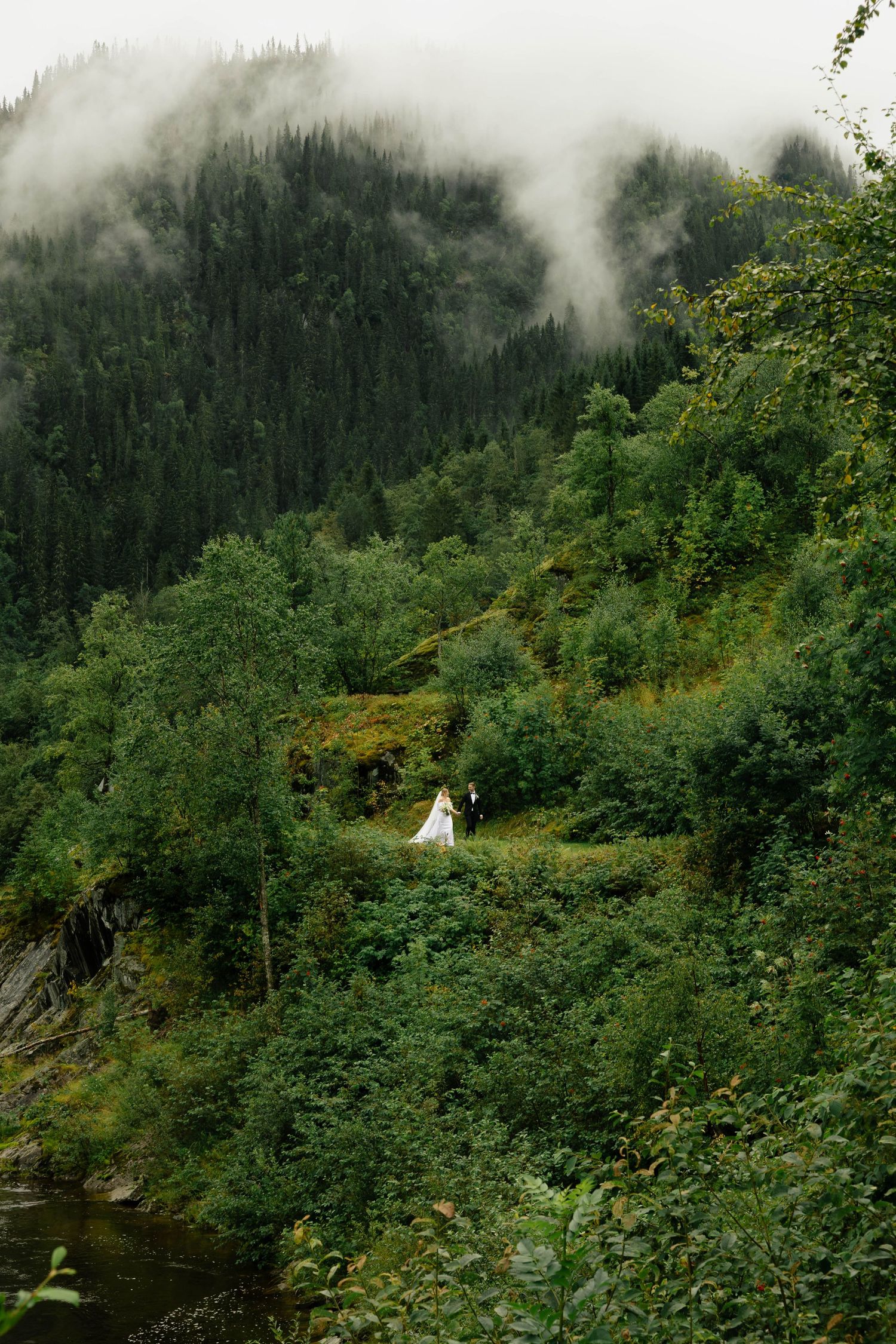 A bride and groom stand on a lush green misty mountain hillside during an outdoor wedding.