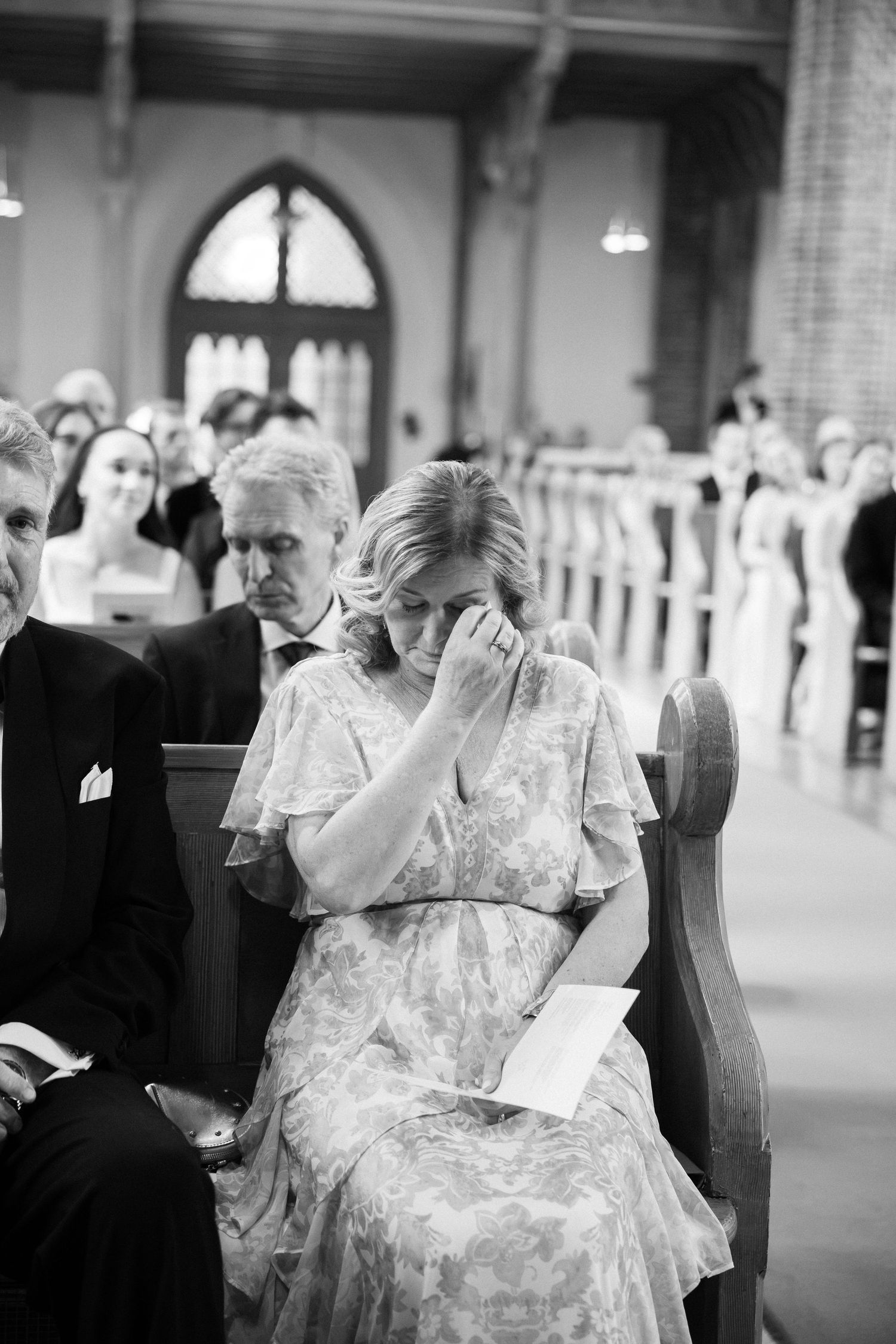 Emotional wedding guest wipes tears while seated in church pew during a heartfelt wedding ceremony.