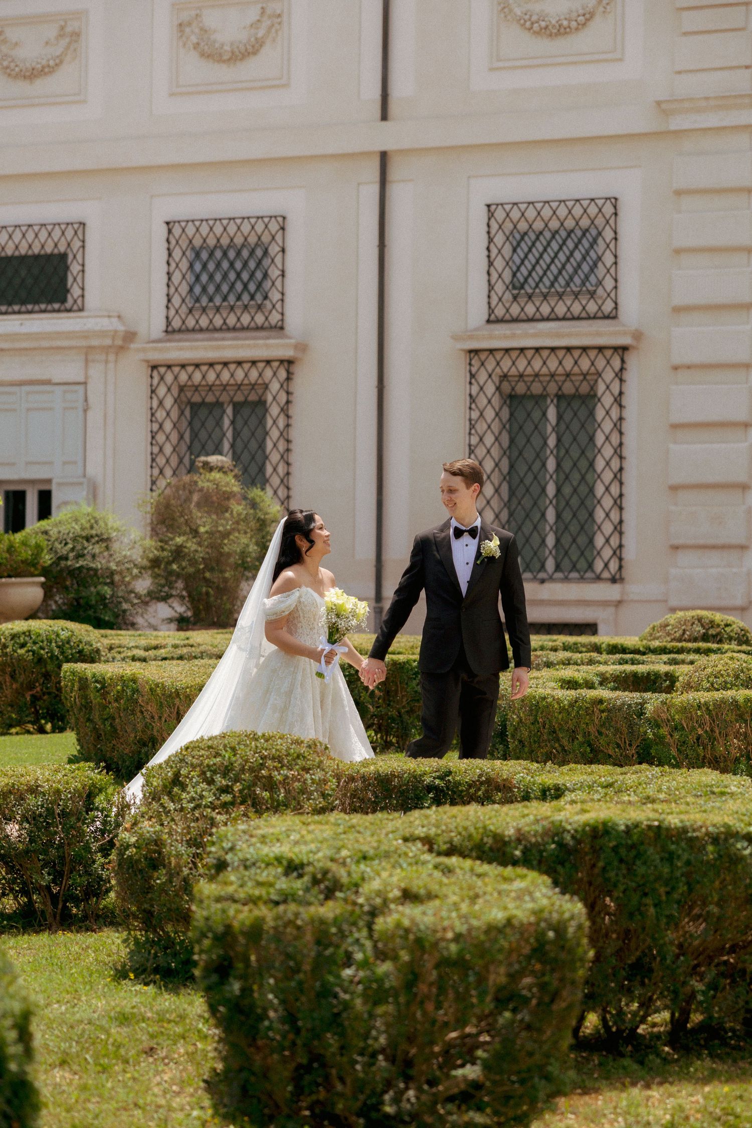 Bride and groom hold hands in a manicured garden outside an elegant villa on their wedding day.