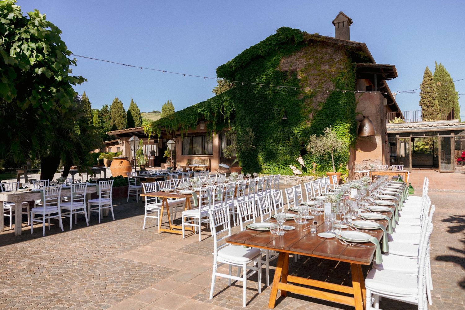 Elegant outdoor wedding reception with white chairs and long tables set in a sunlit Italian villa courtyard surrounded by lush greenery.
