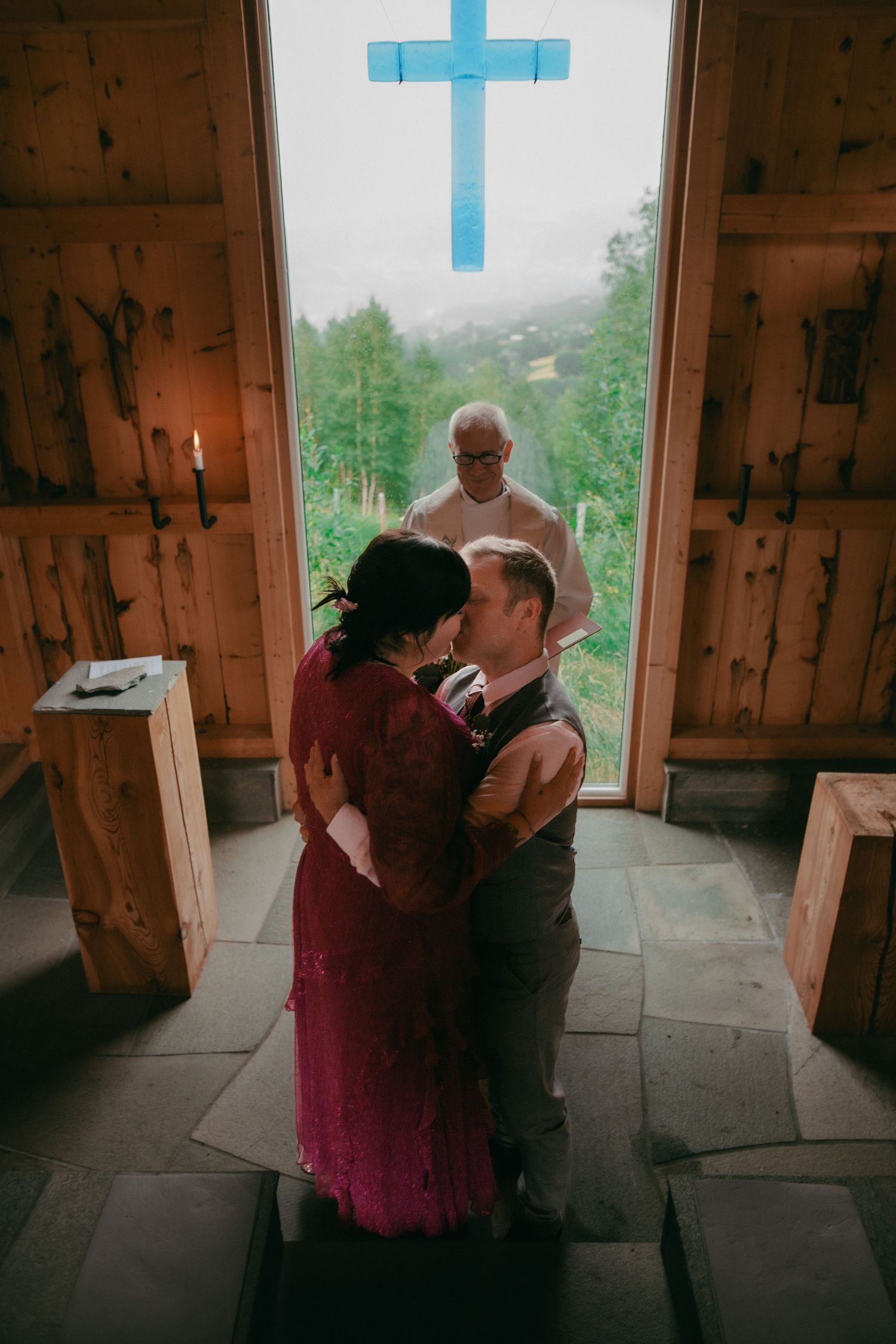Bride and groom sharing first kiss in rustic wooden chapel with blue cross and misty mountain view behind officiant.
