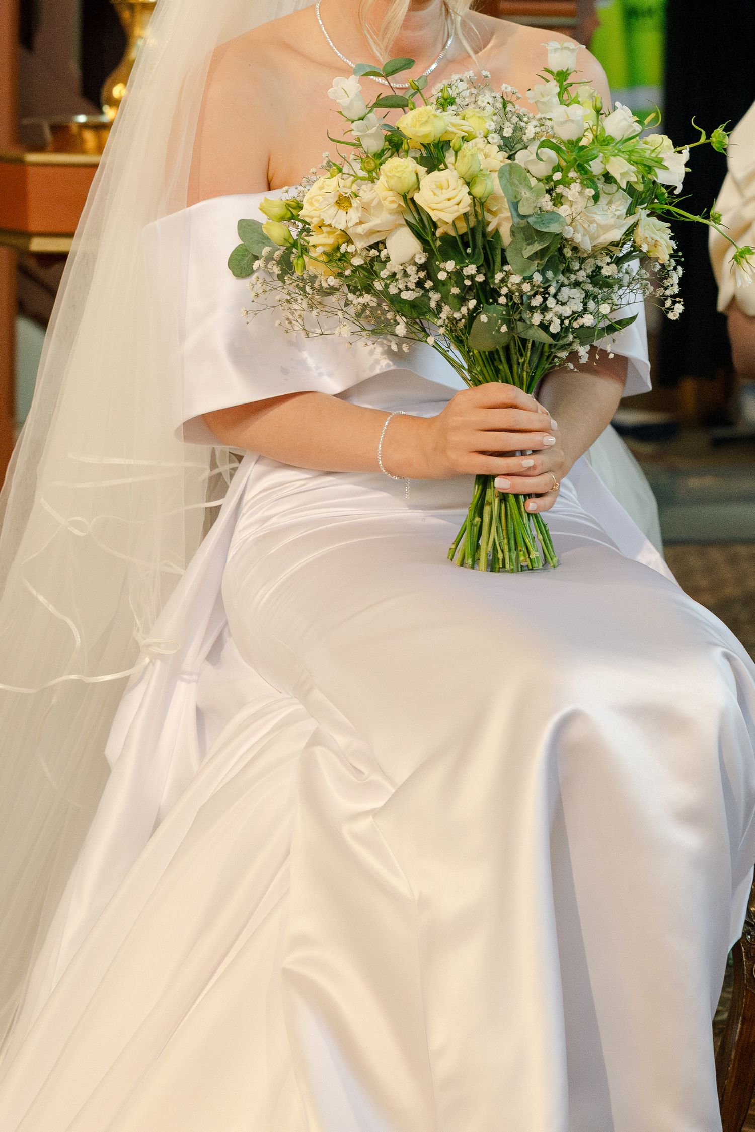 Bride in white satin off-shoulder gown holding elegant bouquet of white roses and gypsophila.