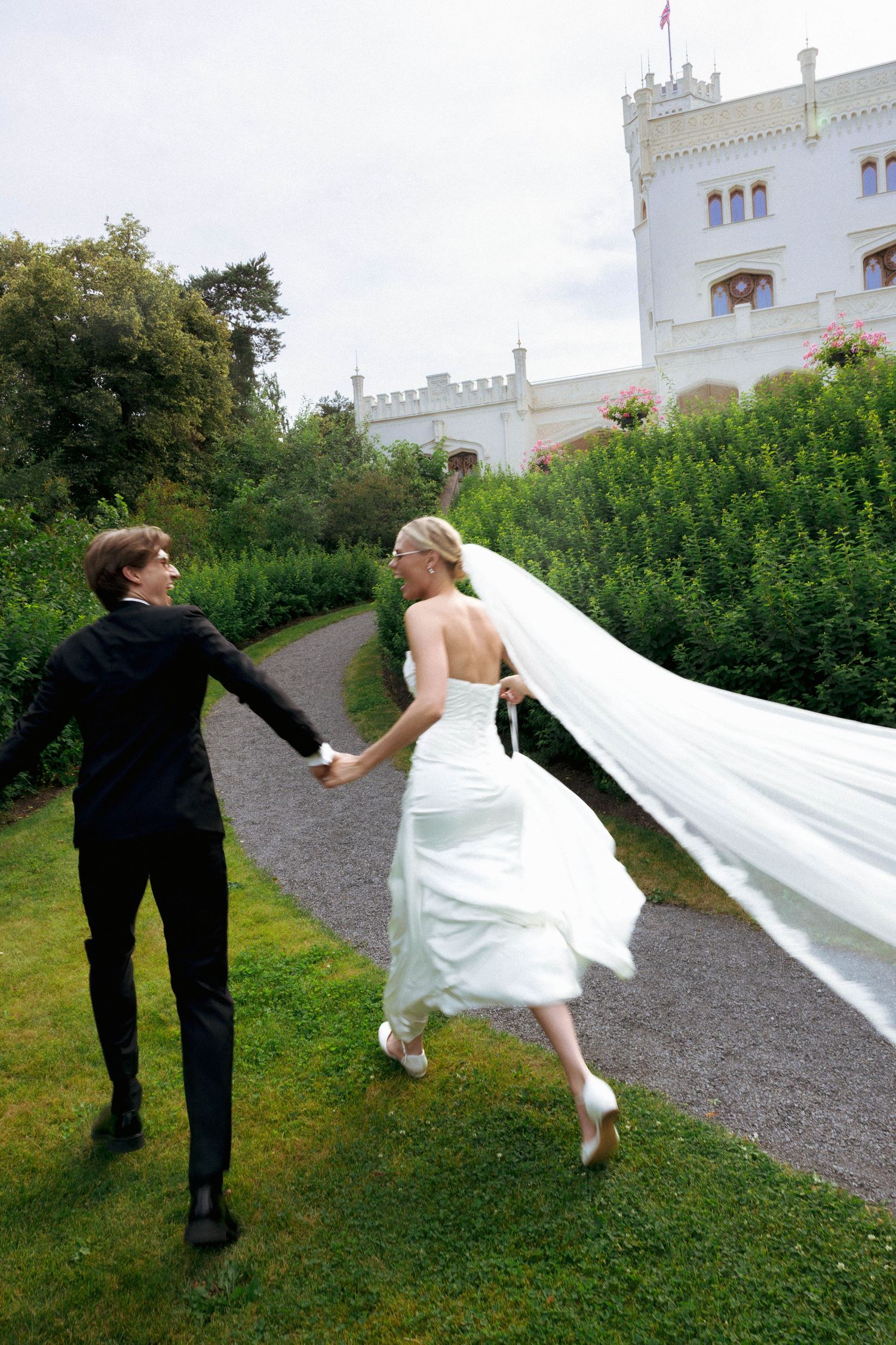 Bride and groom holding hands running toward castle garden path, veil flowing in the wind.