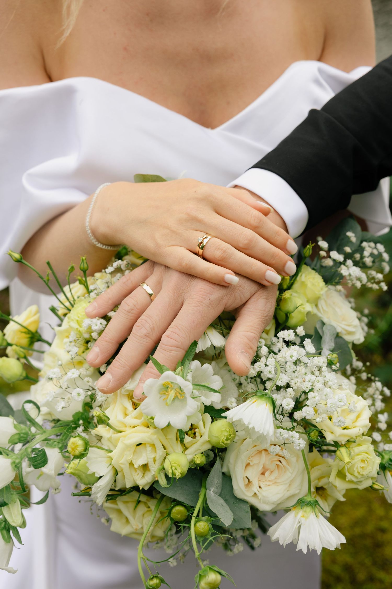 Bride and groom displaying wedding rings on hands resting over a white floral bouquet with roses and baby's breath.