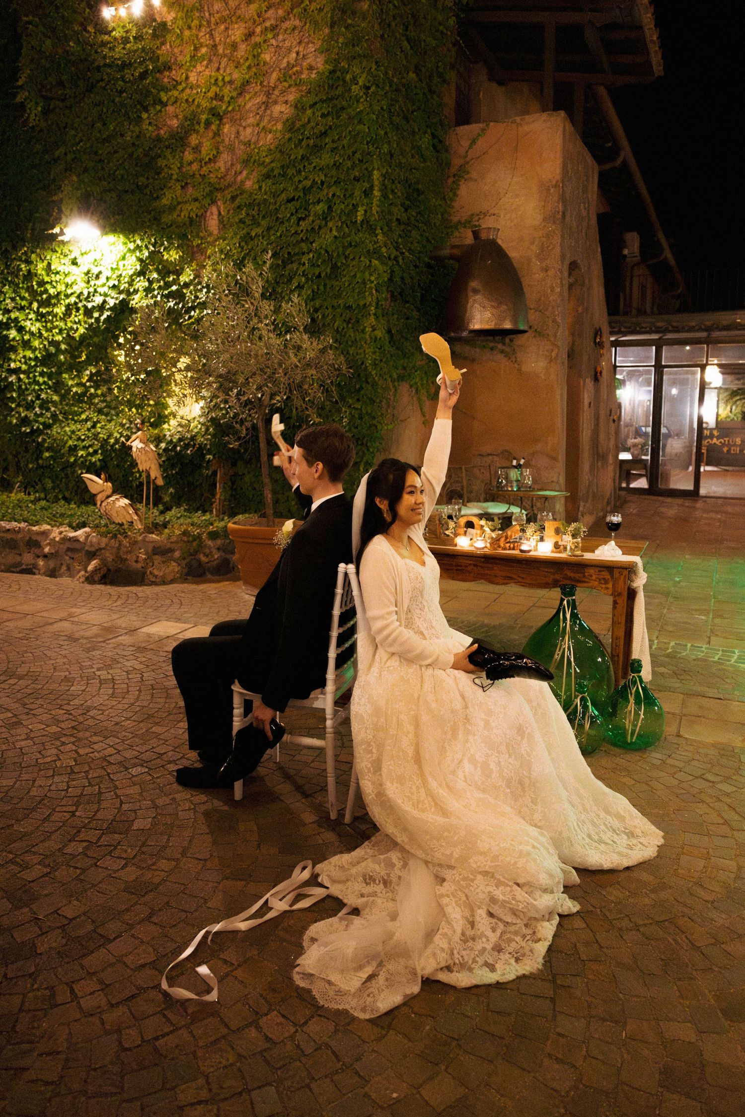 Bride in lace dress raises glass while seated with groom at night outdoor wedding celebration.