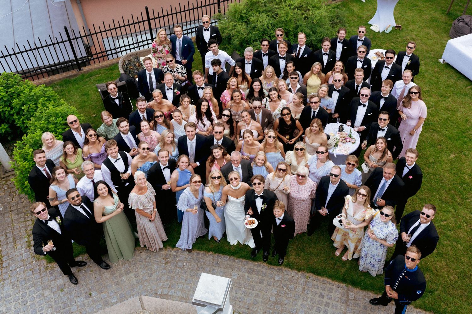 Large wedding group photo from above, guests wearing sunglasses and smiling in sunny outdoor setting.