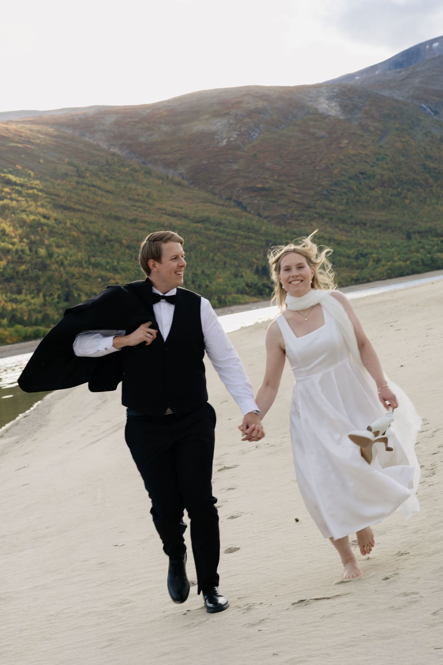 Bride and groom run hand-in-hand on sandy beach with mountain backdrop on their wedding day.