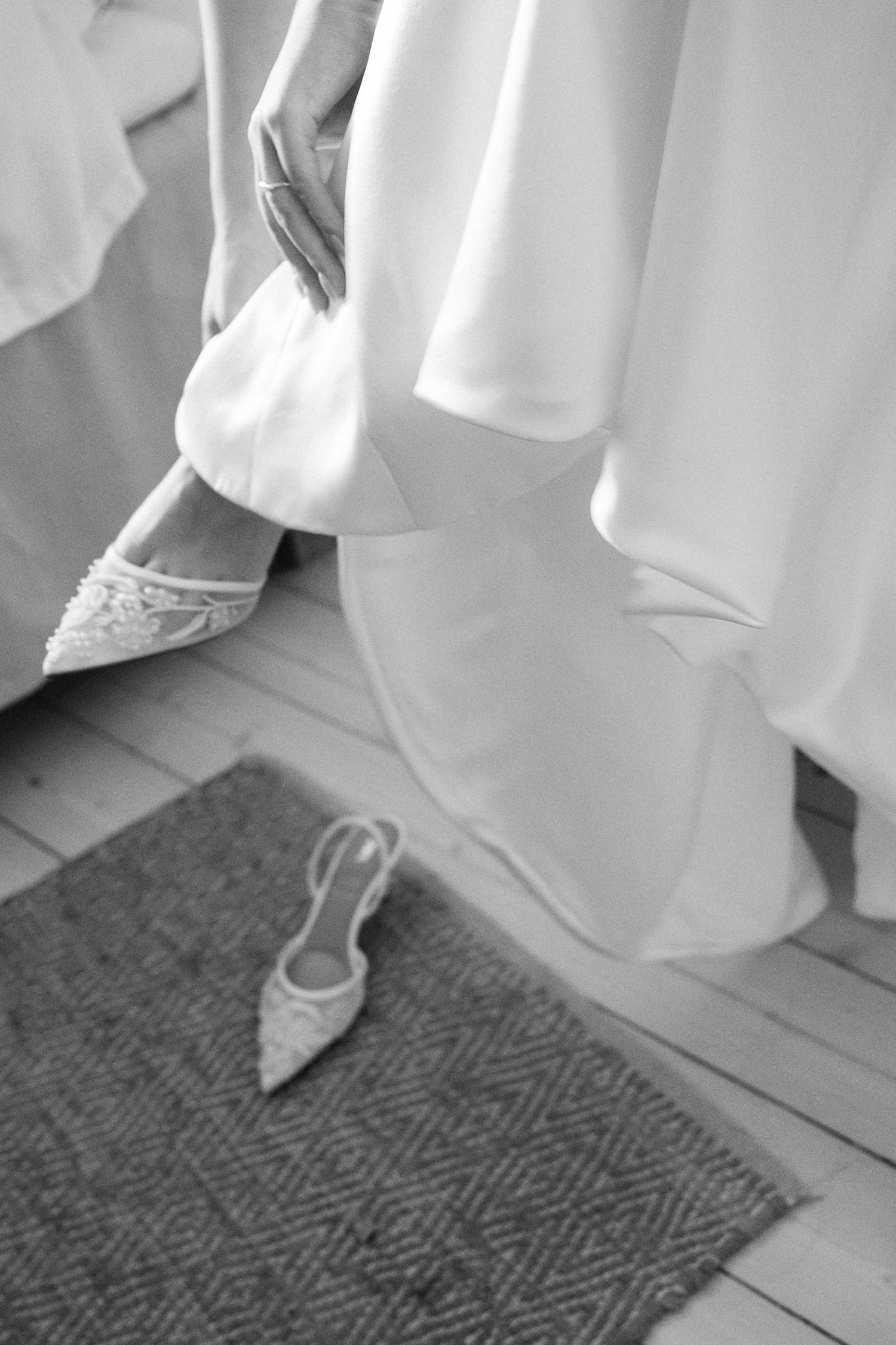 Black and white photo of a bride in a white gown putting on elegant embellished heels on a wooden floor.