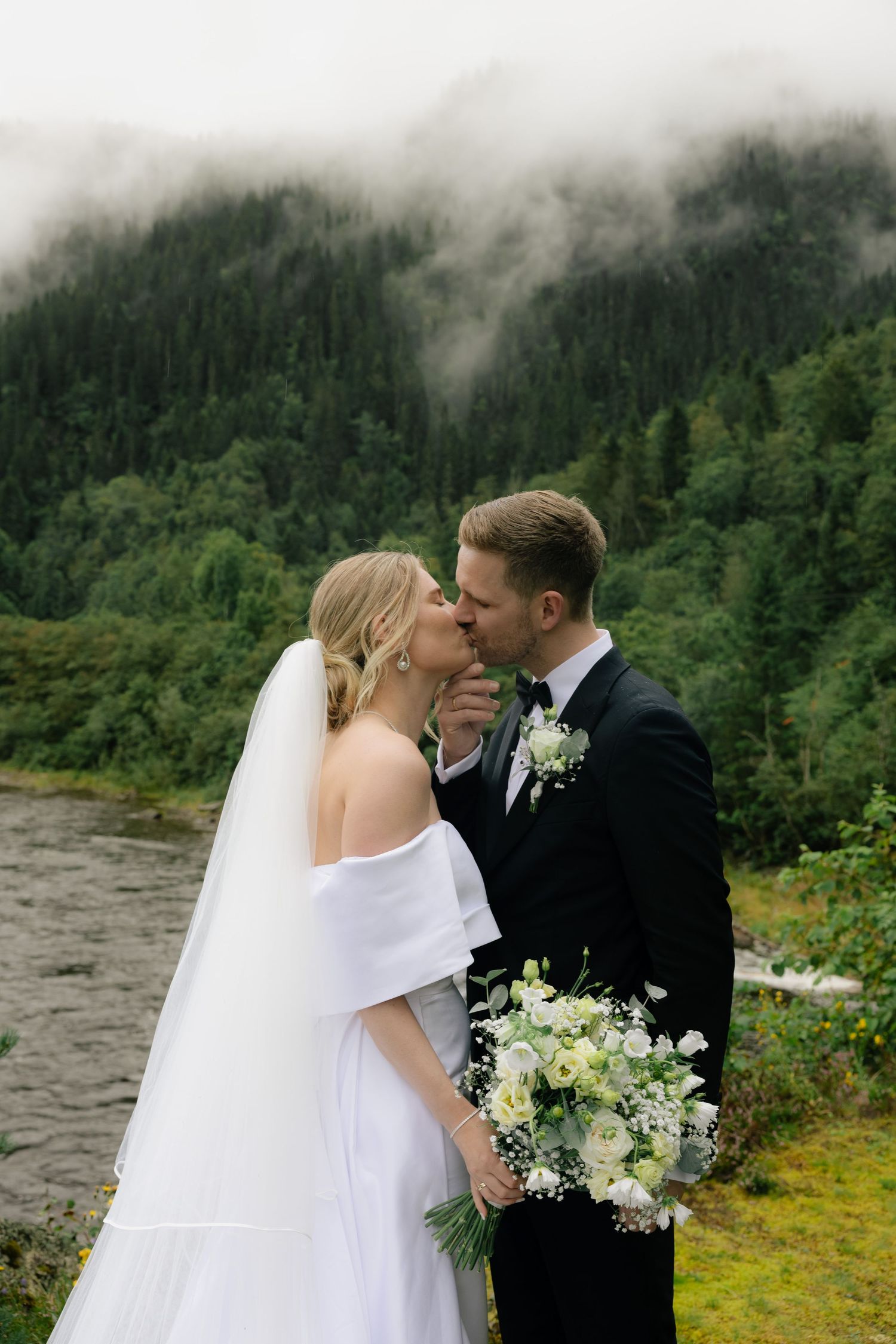 Bride and groom kissing with misty mountain forest backdrop holding white wedding bouquet.
