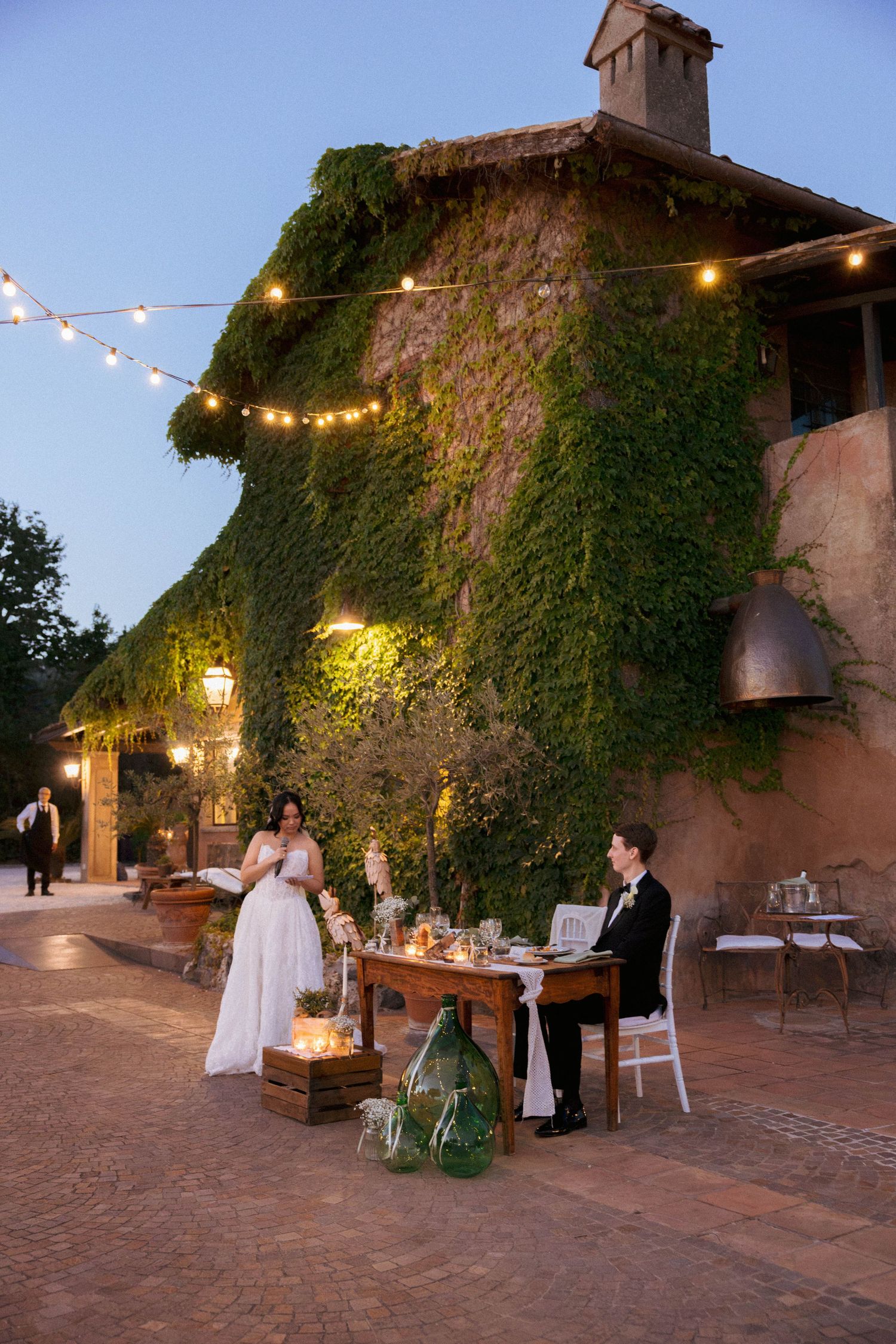 Bride and groom share intimate dinner at rustic ivy-covered venue with warm string lights at dusk.