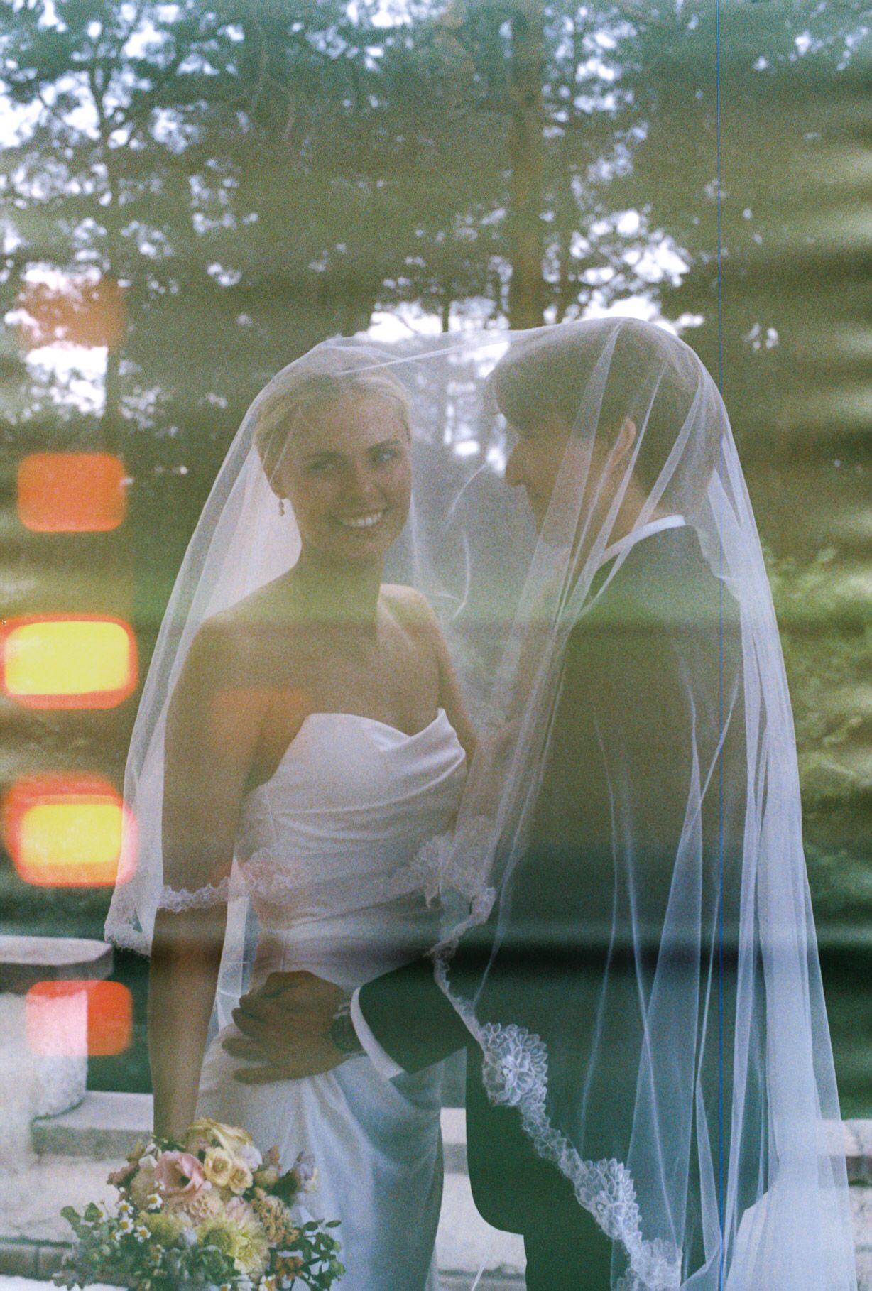 Bride and groom sharing intimate moment under veil, captured through glass reflection with bokeh lights.