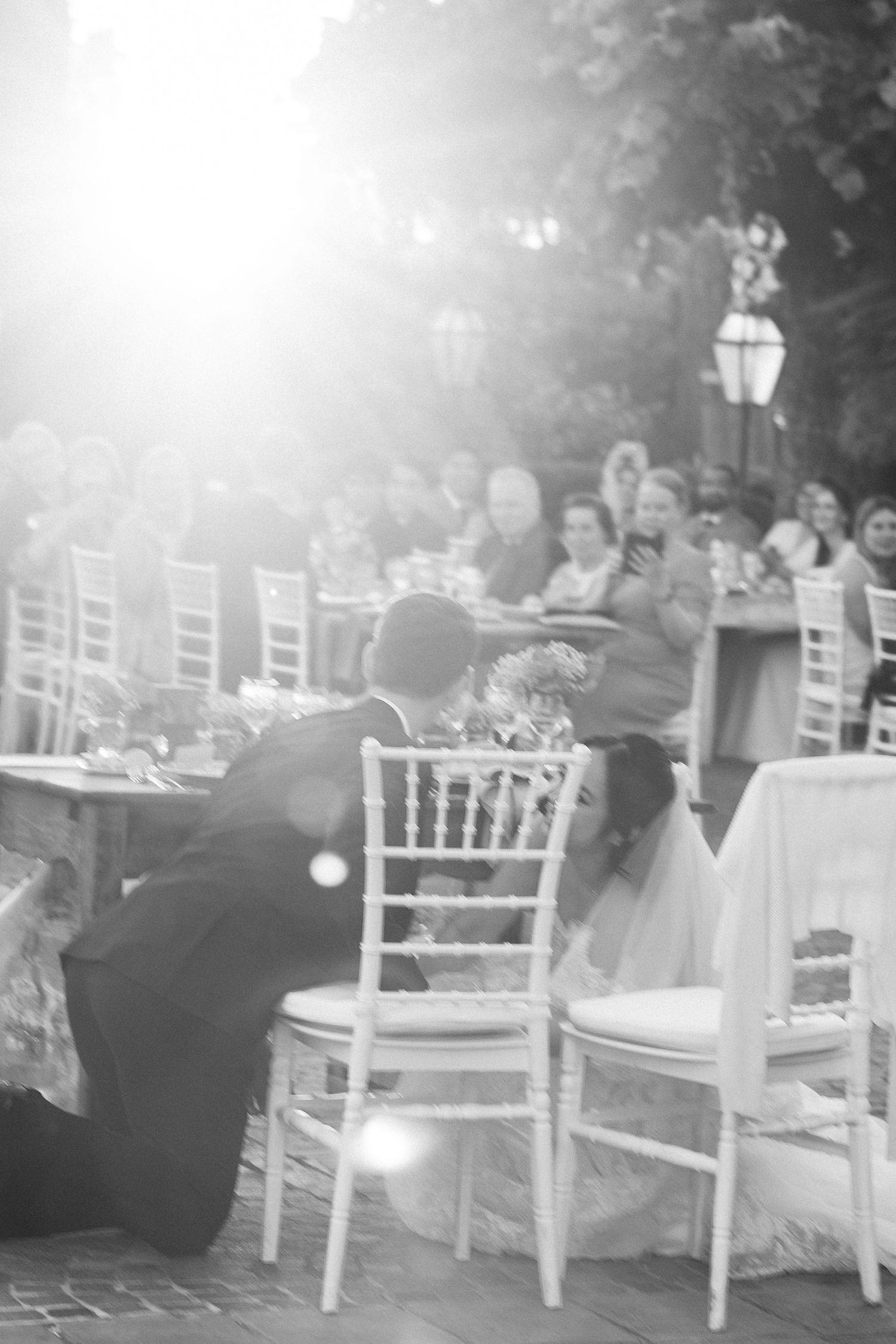 Black and white photo of groom kneeling beside seated bride at outdoor wedding reception.