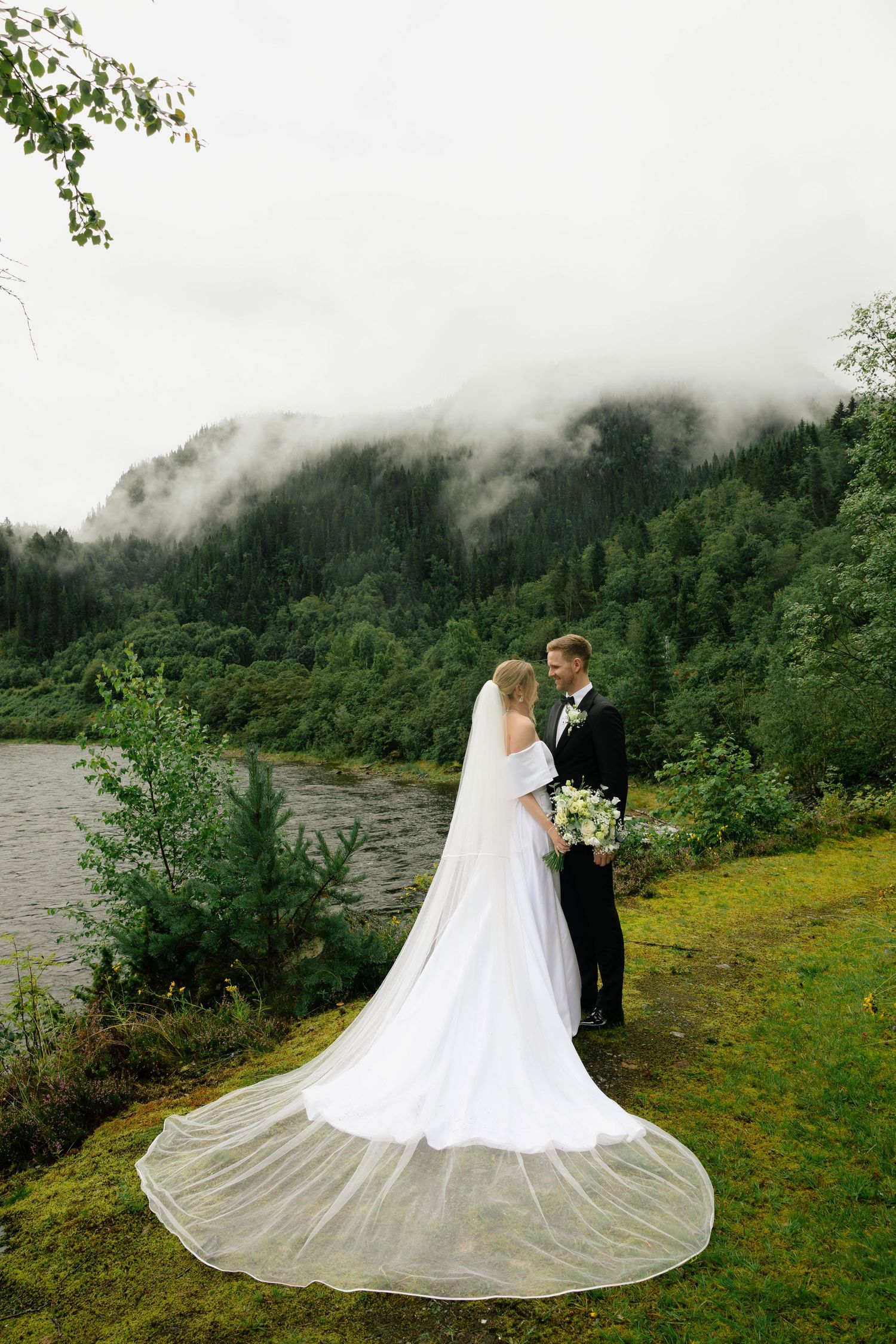 Bride and groom standing by a misty lake surrounded by lush green forest on their wedding day.