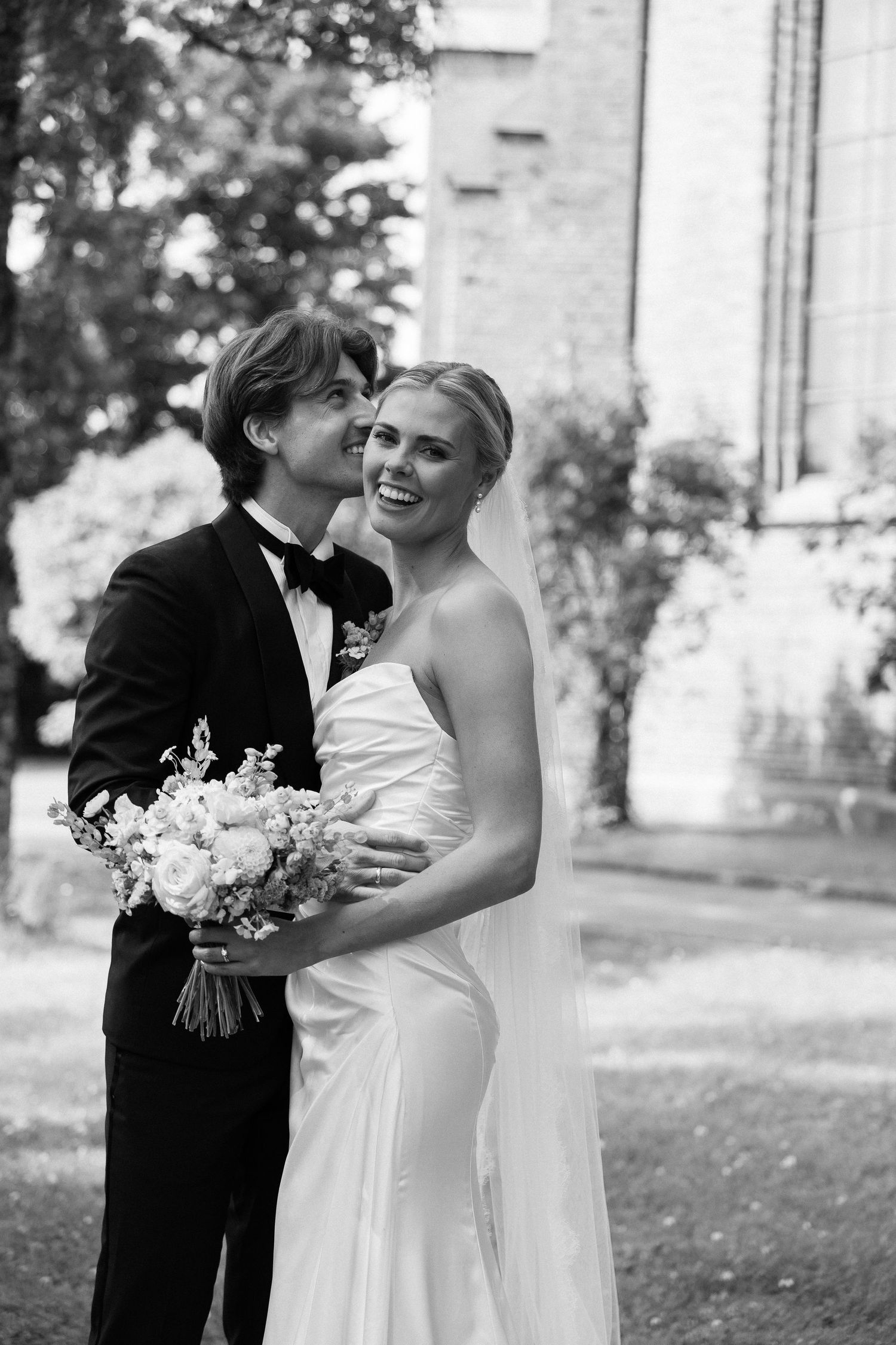 Black and white photo of smiling bride in strapless gown with veil and groom in tuxedo embracing outdoors.