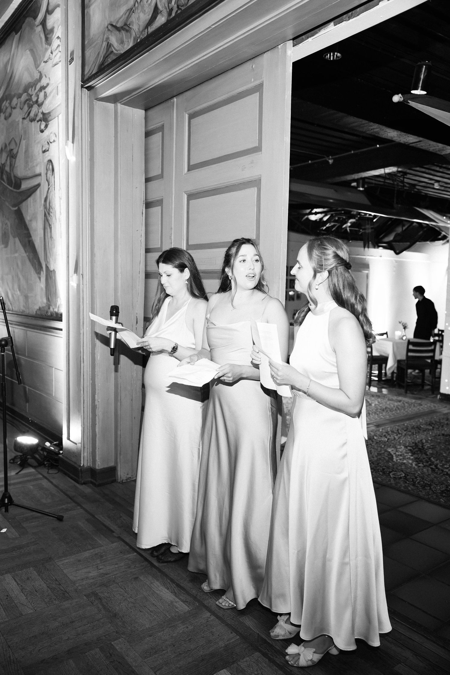 Three bridesmaids in matching satin gowns singing or reading at wedding reception venue.