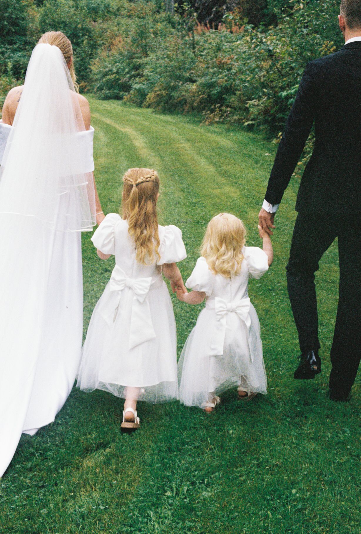 Bride and groom walk hand in hand with two young flower girls in white dresses across a green lawn.