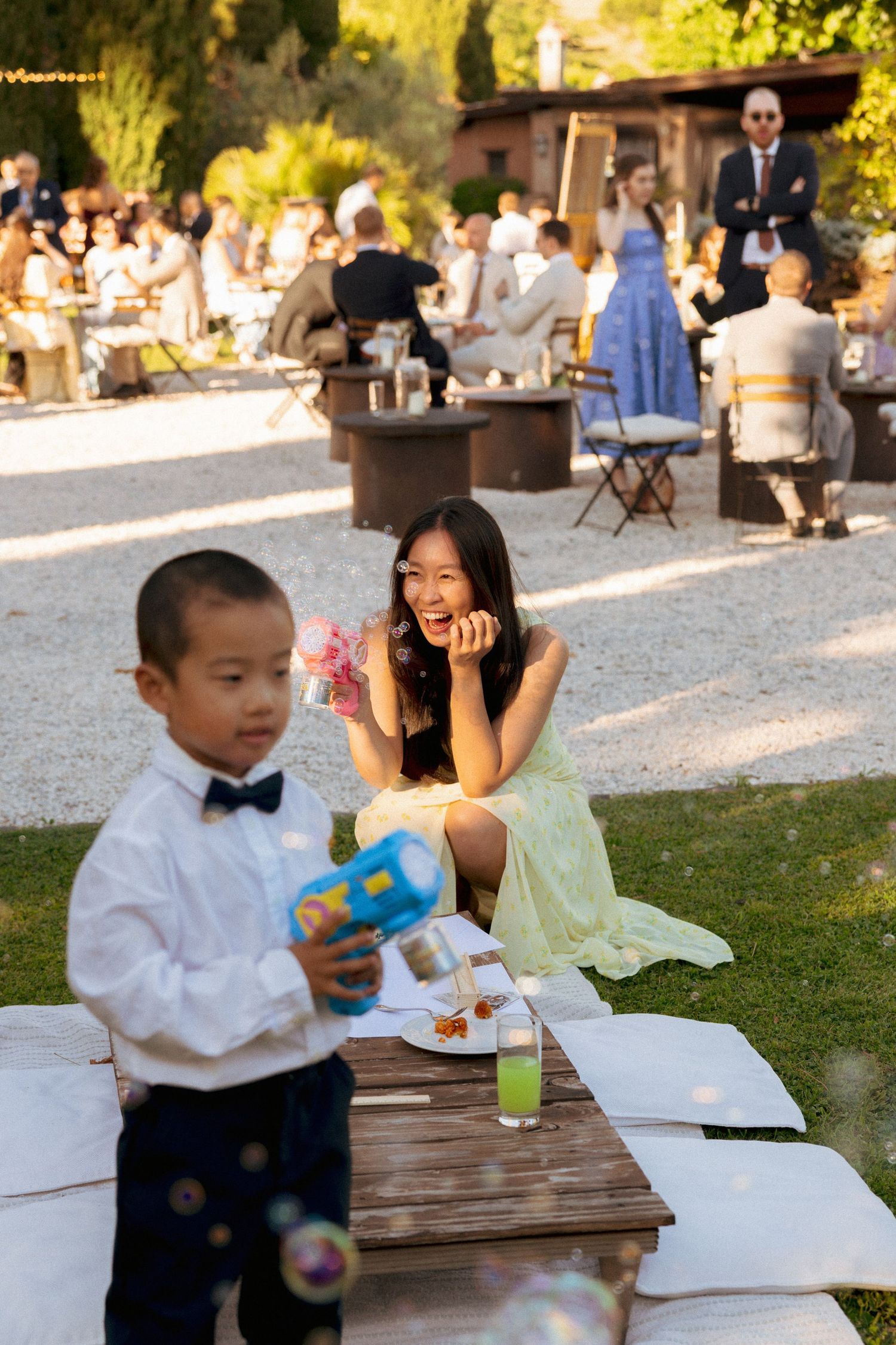 A young boy blows bubbles at an outdoor wedding reception while a smiling woman in a yellow dress watches with delight.