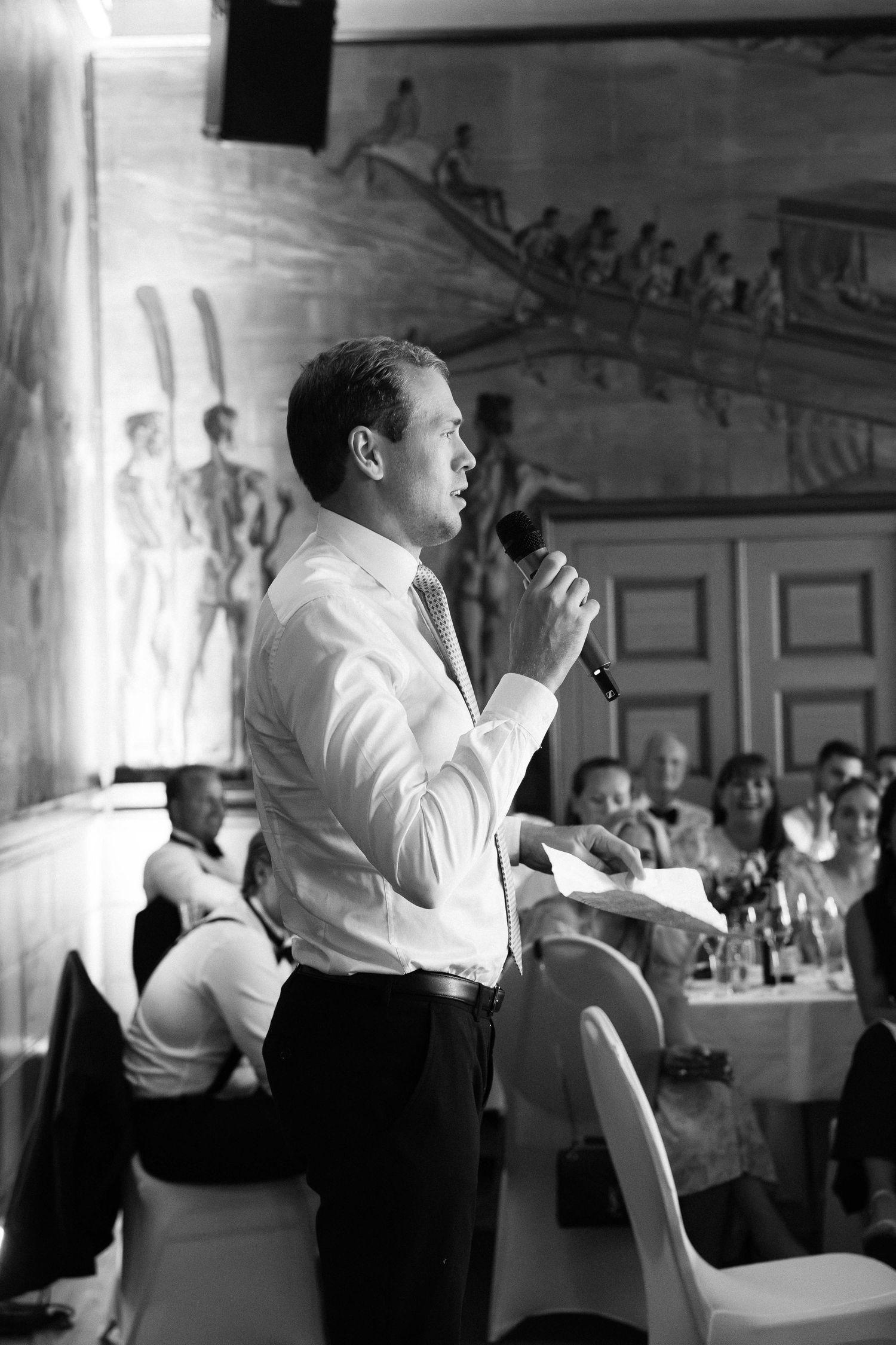Black and white photo of man giving speech at wedding reception holding microphone and notes.
