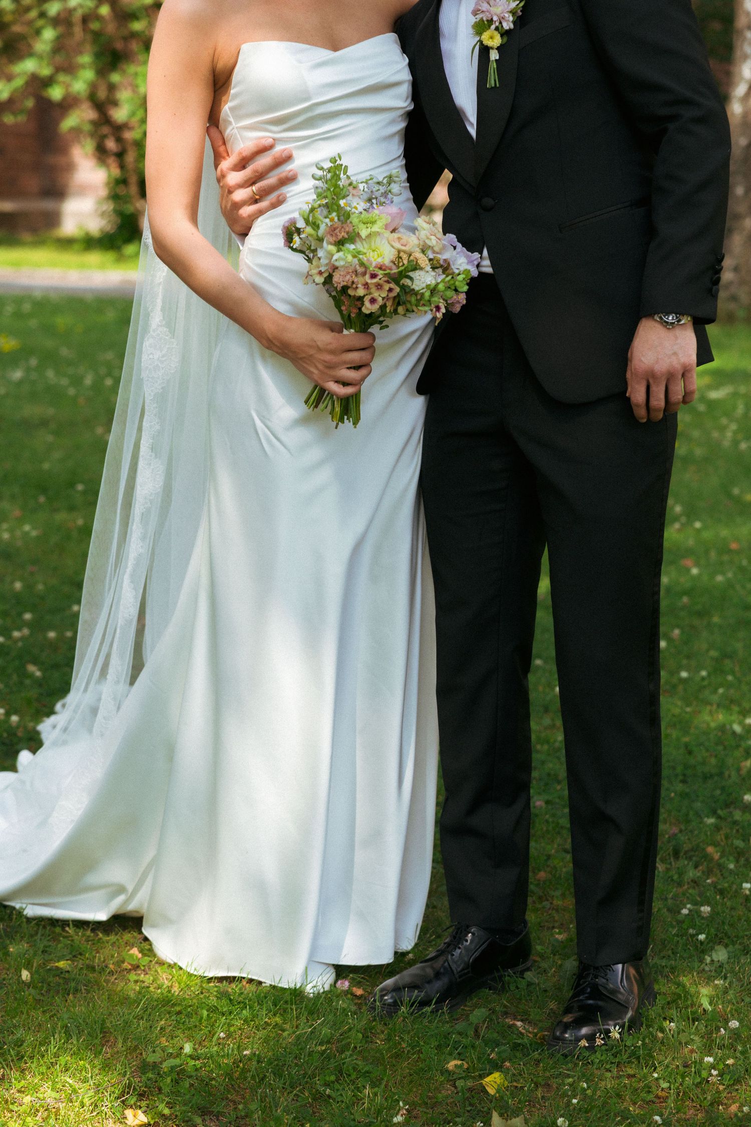 Bride in white strapless gown holding floral bouquet standing with groom in black tuxedo outdoors.