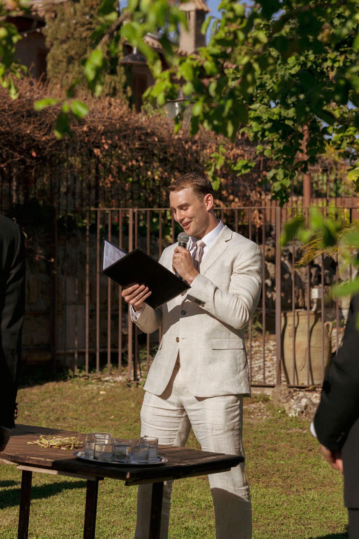 Smiling officiant in beige suit holds microphone and black folder at outdoor wedding ceremony.